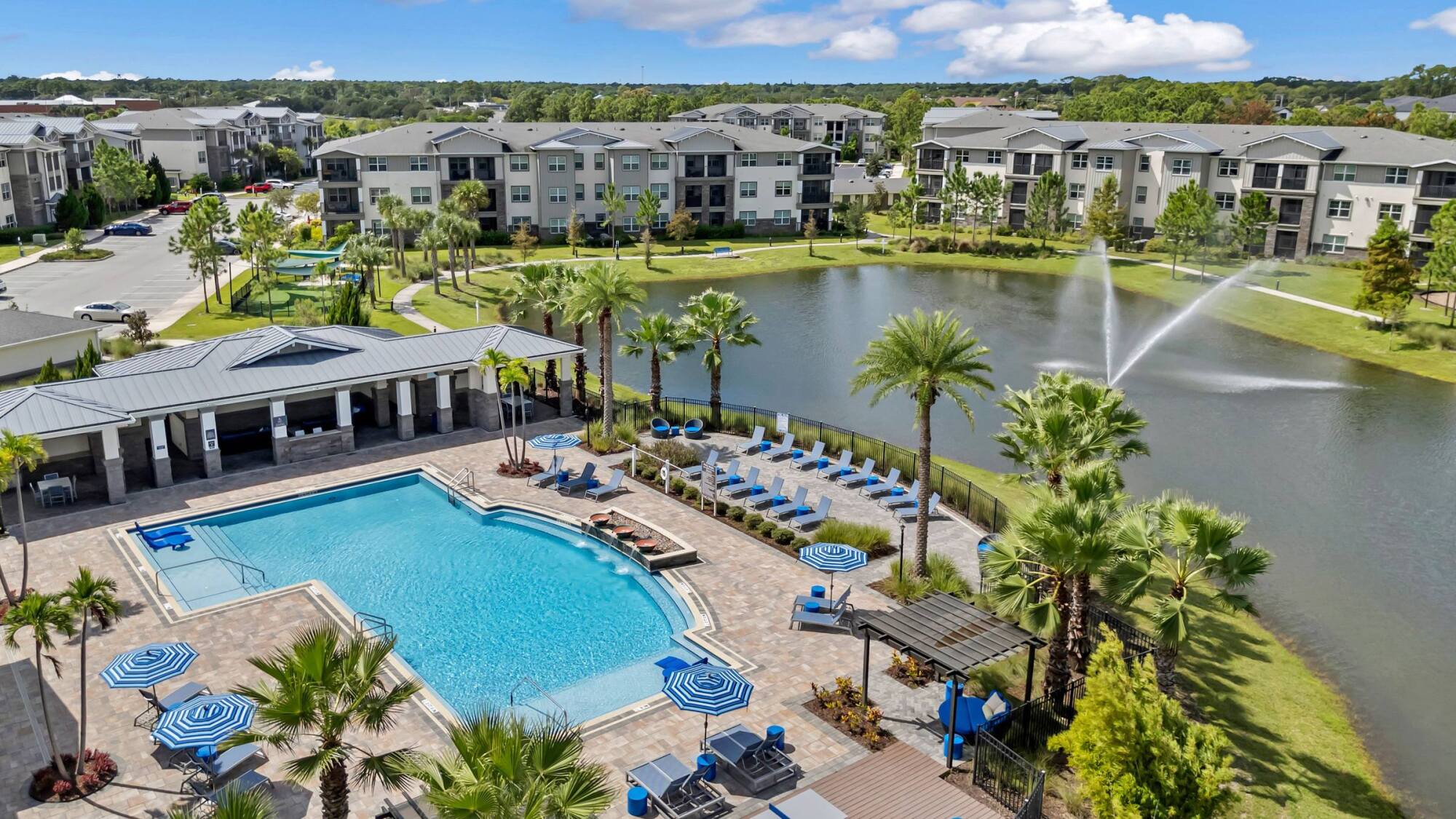 Aerial view of an apartment complex with a pool, lounge chairs, palm trees, and a pond with a fountain.