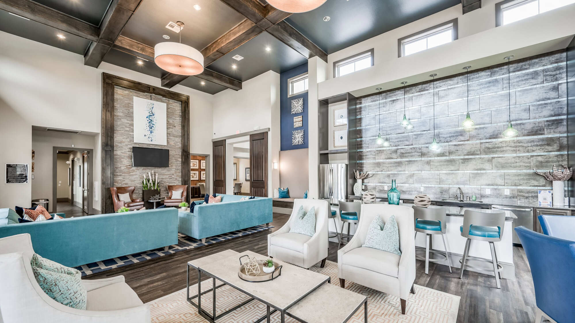 Upscale clubhouse lounge with teal sofas, white accent chairs, exposed wood beam ceilings, stone accent wall, drum pendant lights, and a barstool seating area near a textured tile feature wall.