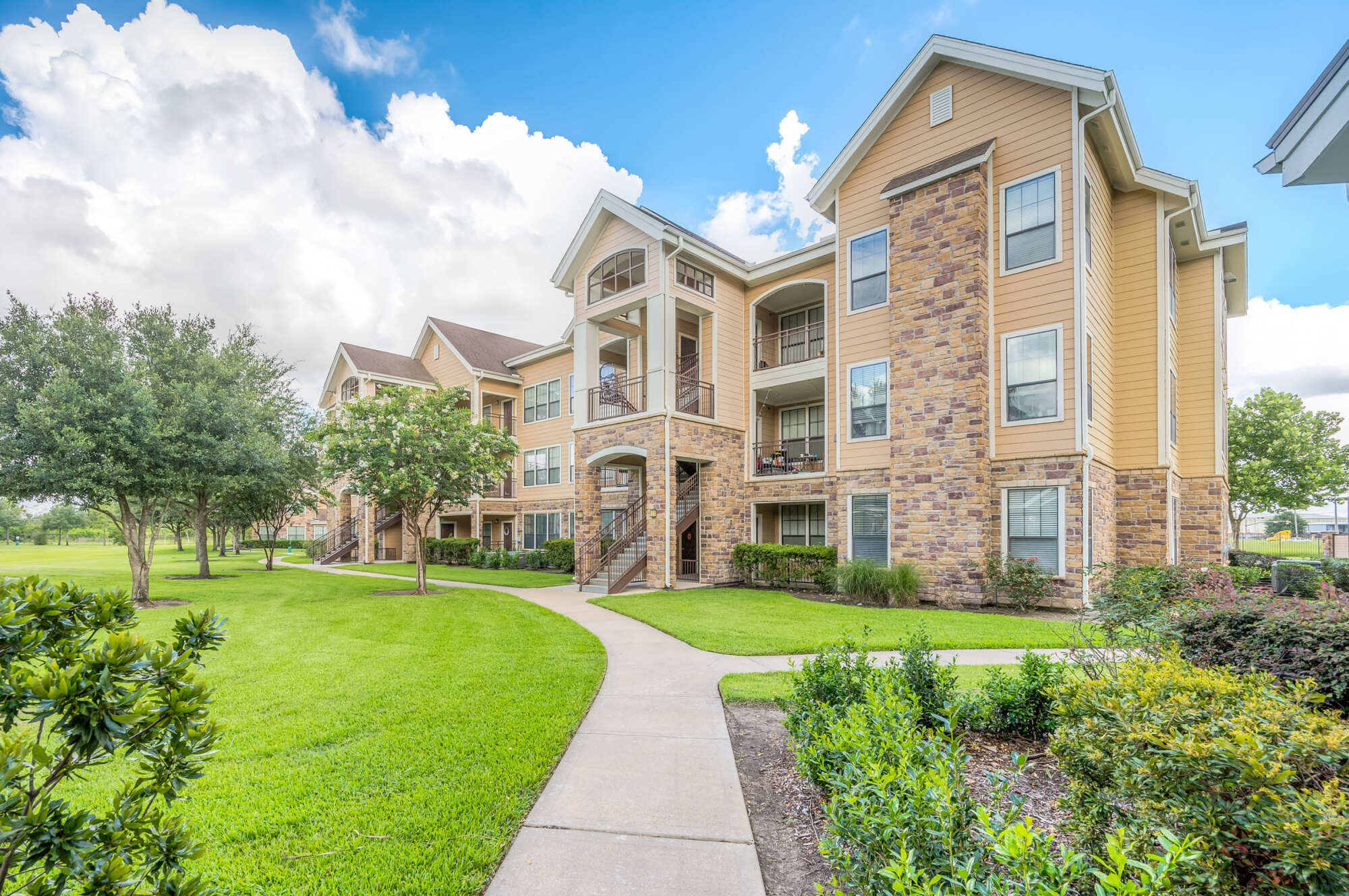 Three-story apartment building with tan siding, stone veneer accents, arched entryway, private balconies, manicured landscaping, and a sidewalk leading toward the entrance.