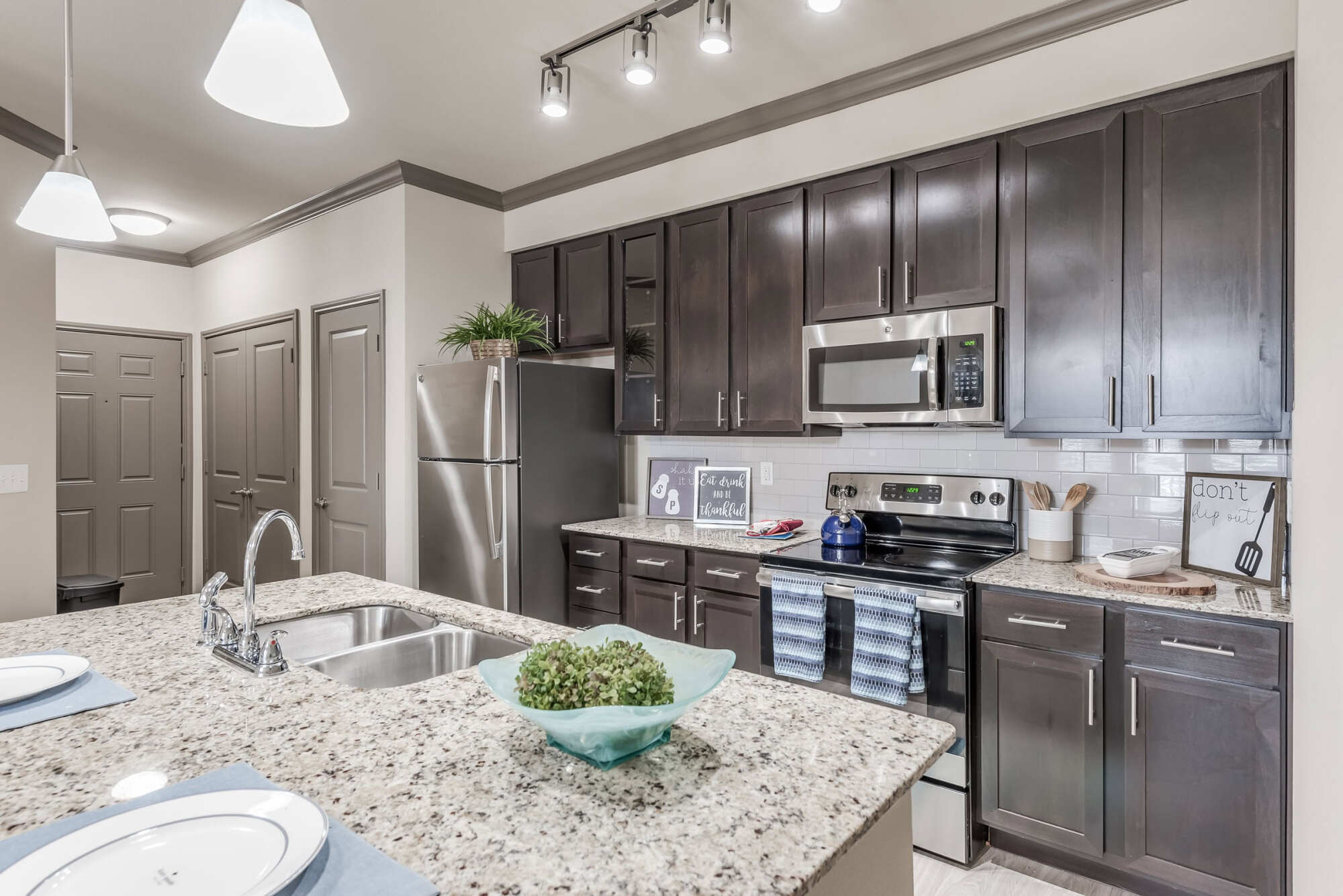 Stylish apartment kitchen with espresso cabinets, granite countertops, stainless steel appliances, white subway tile backsplash, decorative pendant lights, and a center island with double sink.
