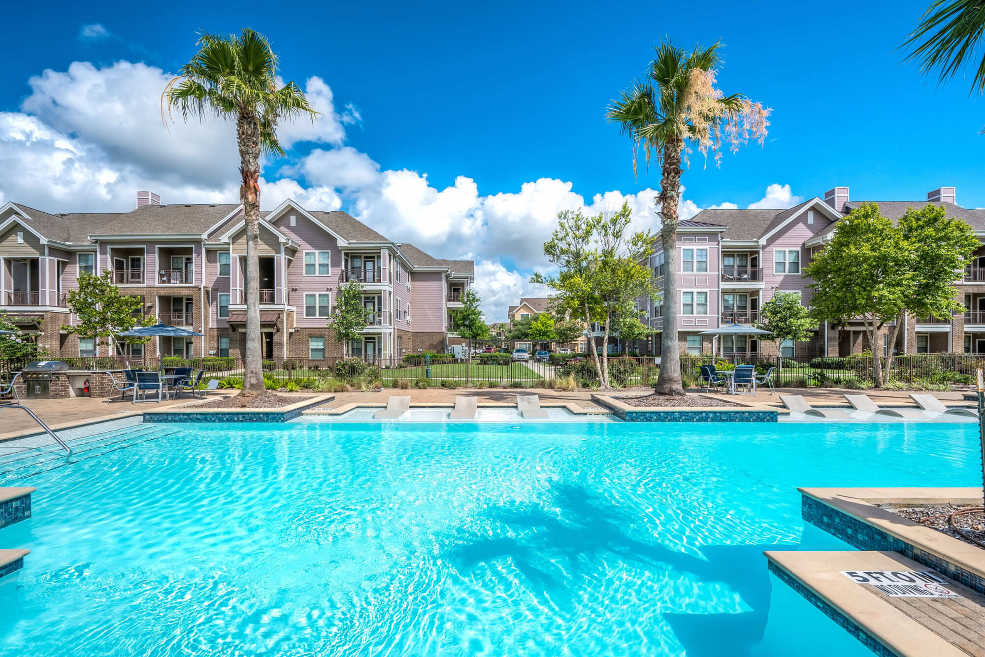 Resort-style swimming pool with tropical palm trees, paver decking, lounge chairs, and pink stucco apartment buildings in the background under a partly cloudy blue sky.