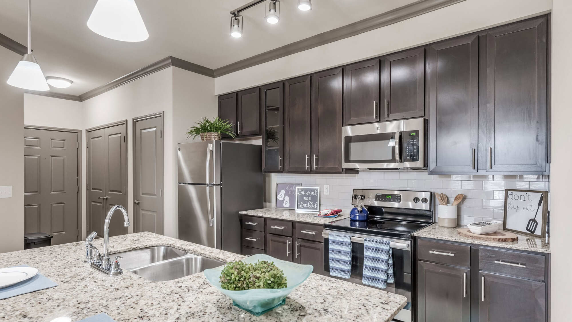 Apartment kitchen featuring dark espresso cabinetry, granite countertops, stainless steel appliances, white subway tile backsplash, modern pendant and track lighting, and farmhouse decor accents.