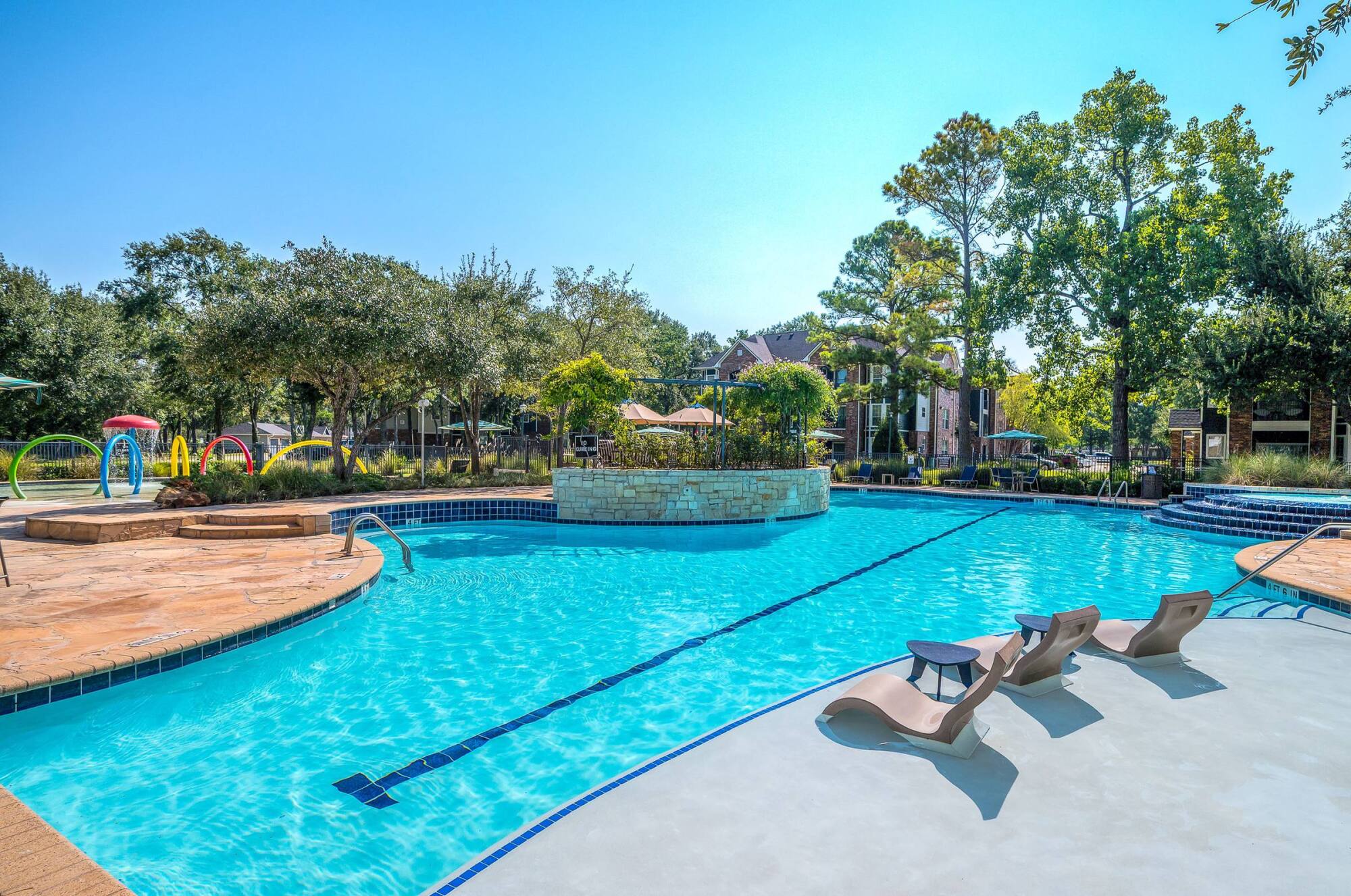 Outdoor swimming pool with lounge chairs in shallow water, surrounded by trees and apartment buildings.