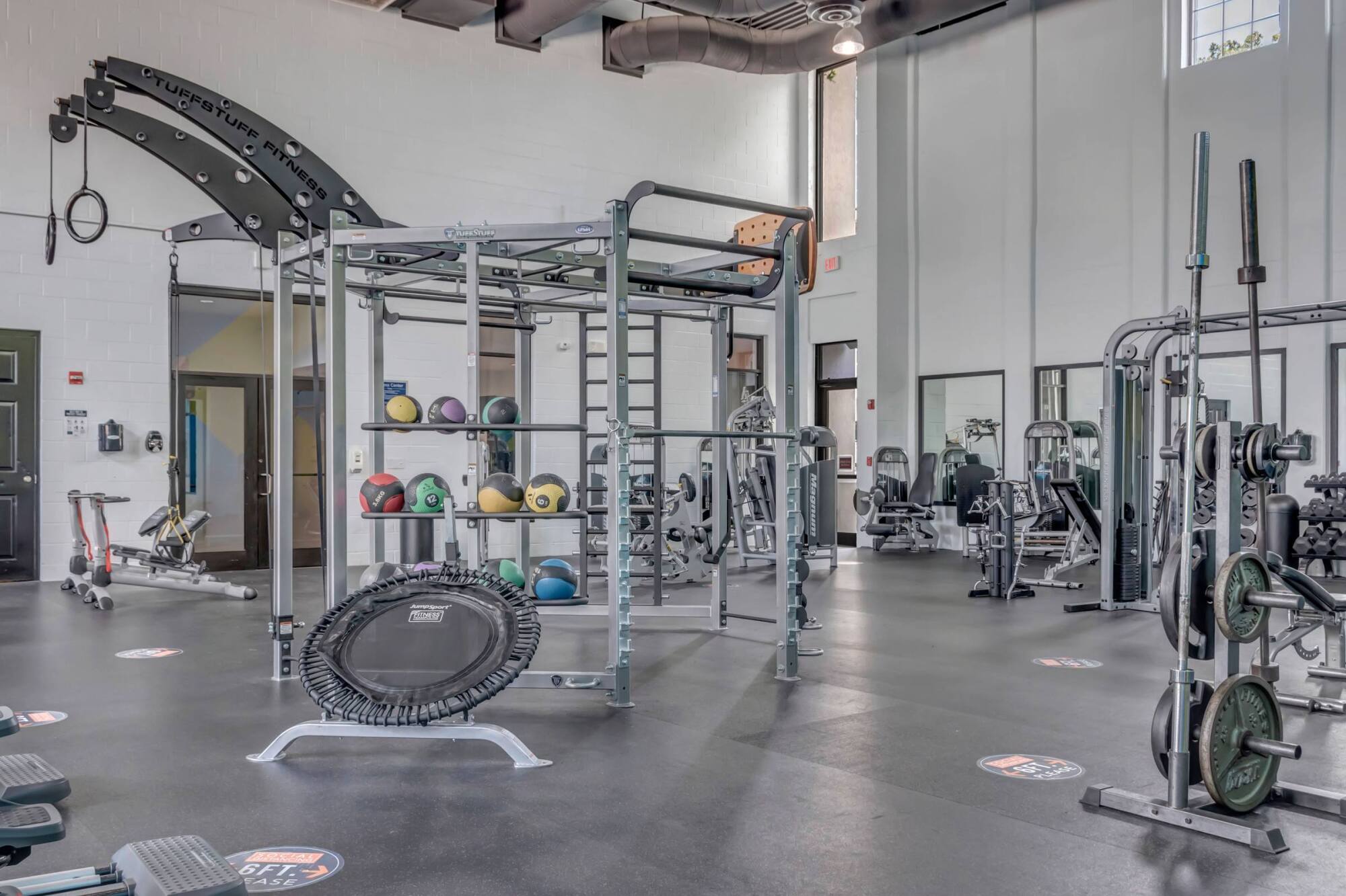 Spacious gym with workout equipment, medicine balls, weights, a trampoline, and high windows letting in light.