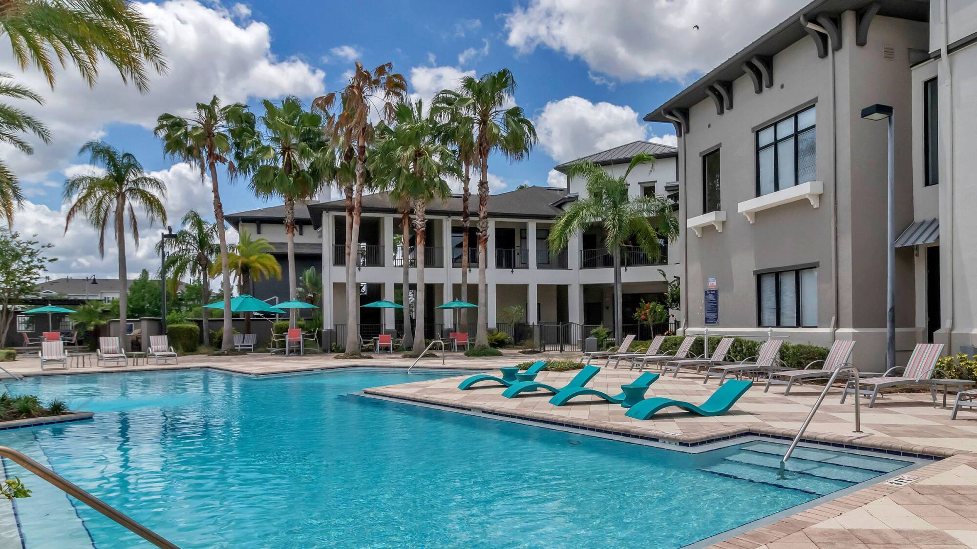Resort-style outdoor pool with lounge chairs, palm trees, and modern apartment buildings under a blue sky.