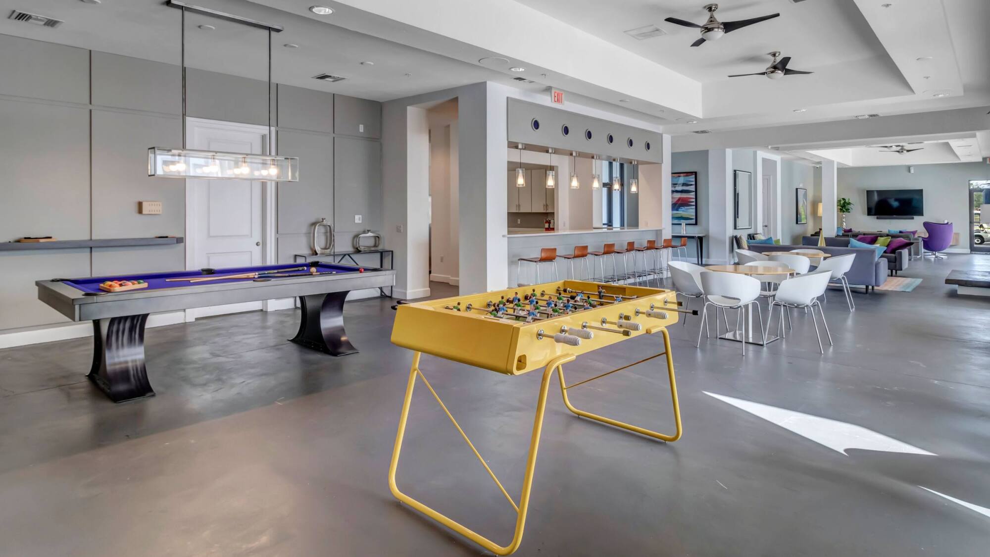 Modern game room with a yellow foosball table, pool table, bar seating, and lounge area in the background.