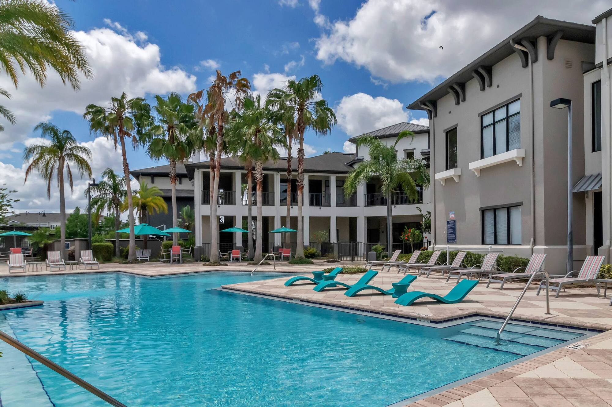 Resort-style apartment pool with lounge chairs, palm trees, and umbrellas under a partly cloudy sky.