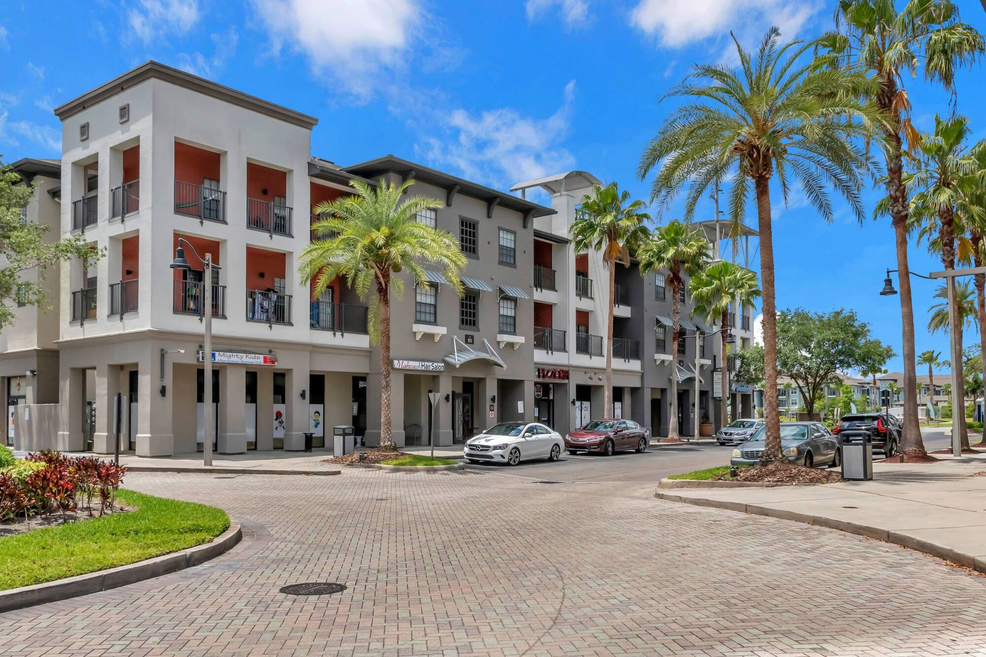 Modern apartment buildings with palm trees and parked cars on a sunny day in an urban area.