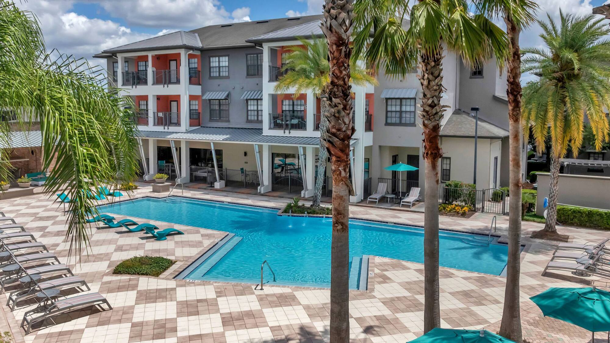 Outdoor swimming pool with lounge chairs, palm trees, and a modern apartment building in the background.