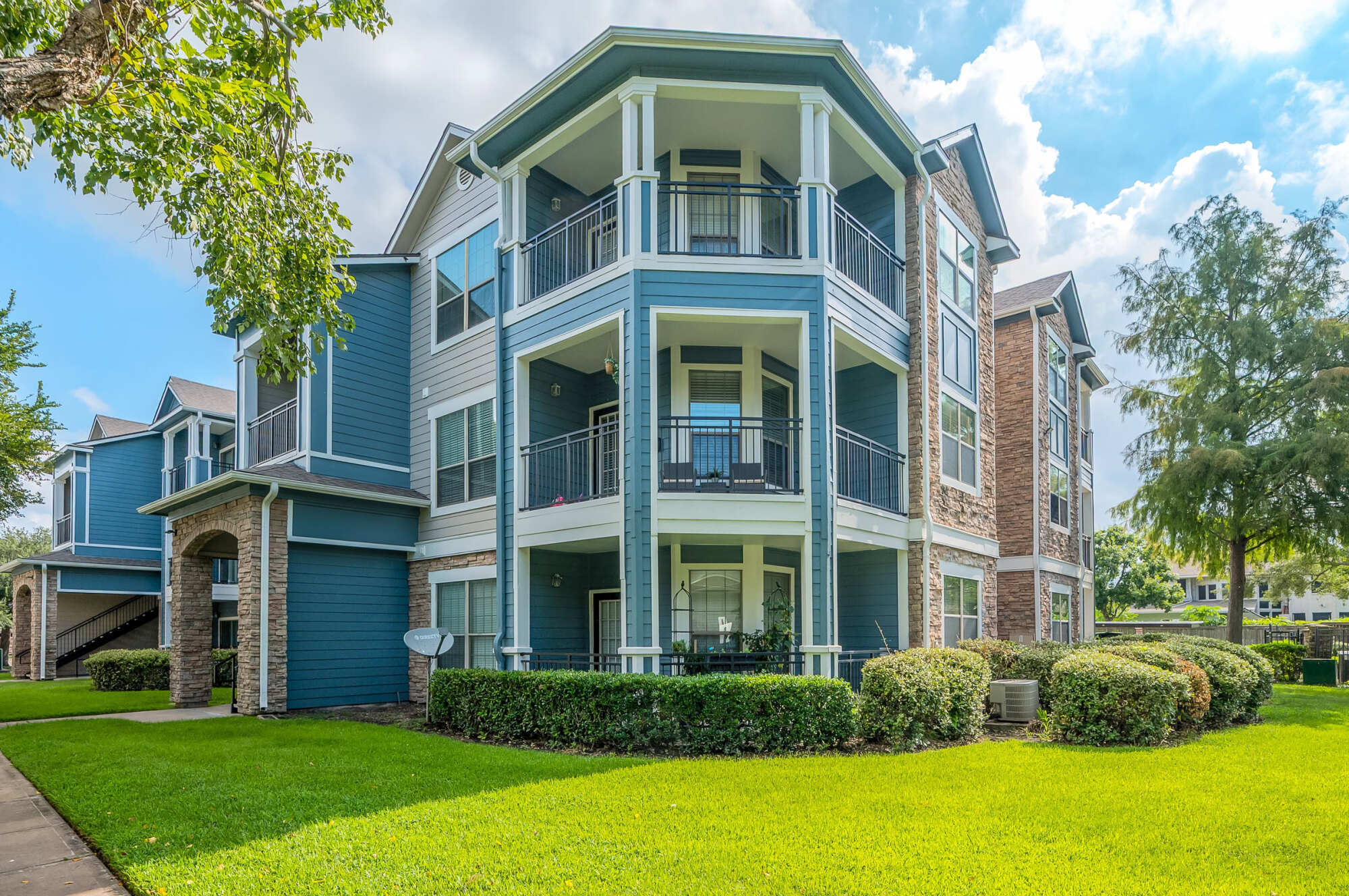 Three-story teal blue apartment building with white columns, stacked private balconies, stone veneer accents at the base, a manicured lawn, and partly cloudy skies overhead.