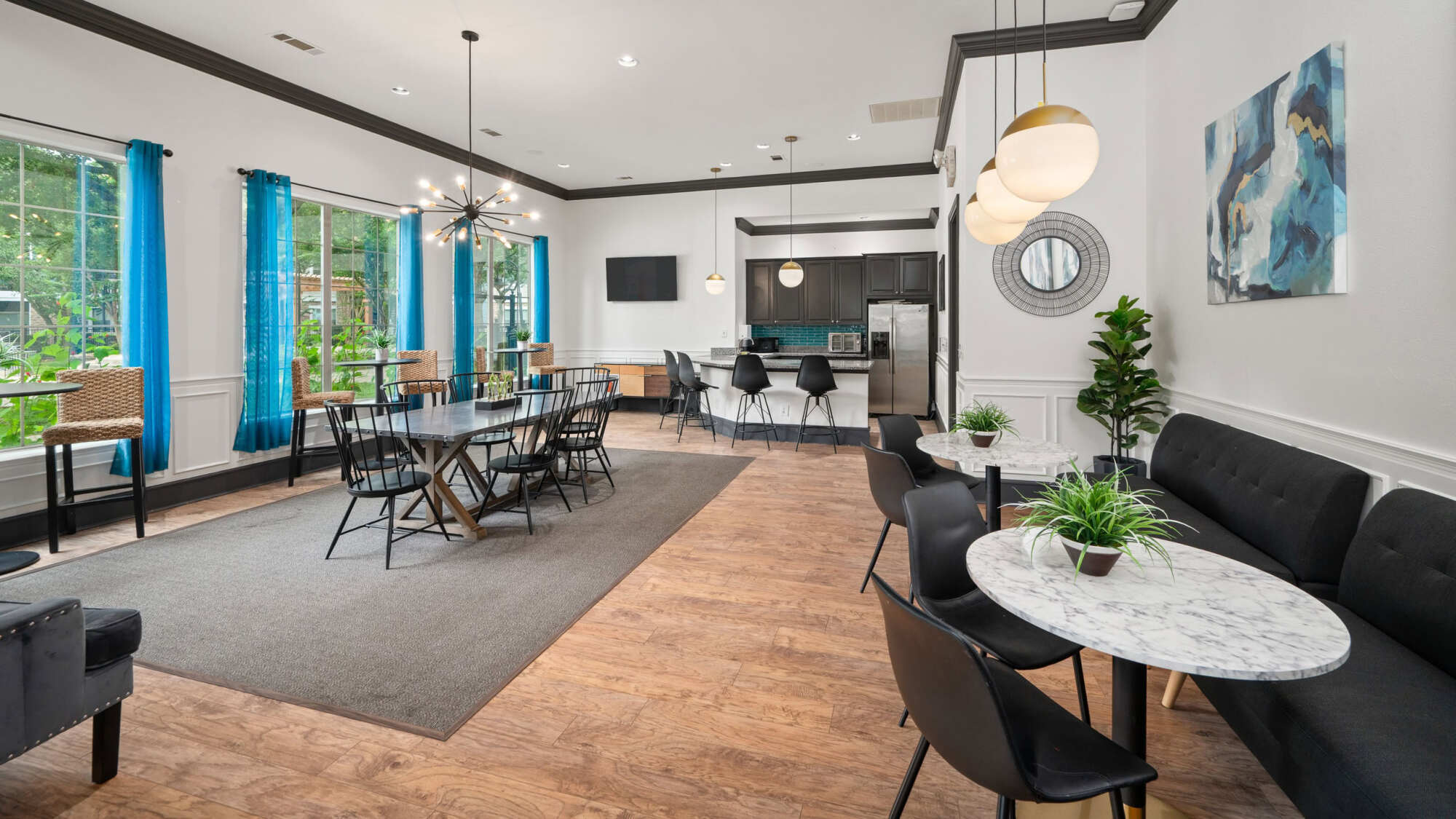 Stylish clubhouse lounge featuring a long communal table, modern chandelier lighting, dark accents, abstract wall art, an oval marble bistro table, and a kitchen counter in the background.