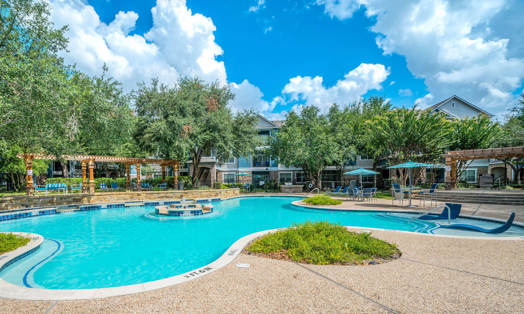 Curving resort-style pool surrounded by paver decking, lounge chairs, blue umbrellas, a wooden pergola, lush trees, and apartment buildings under a partly cloudy sky.