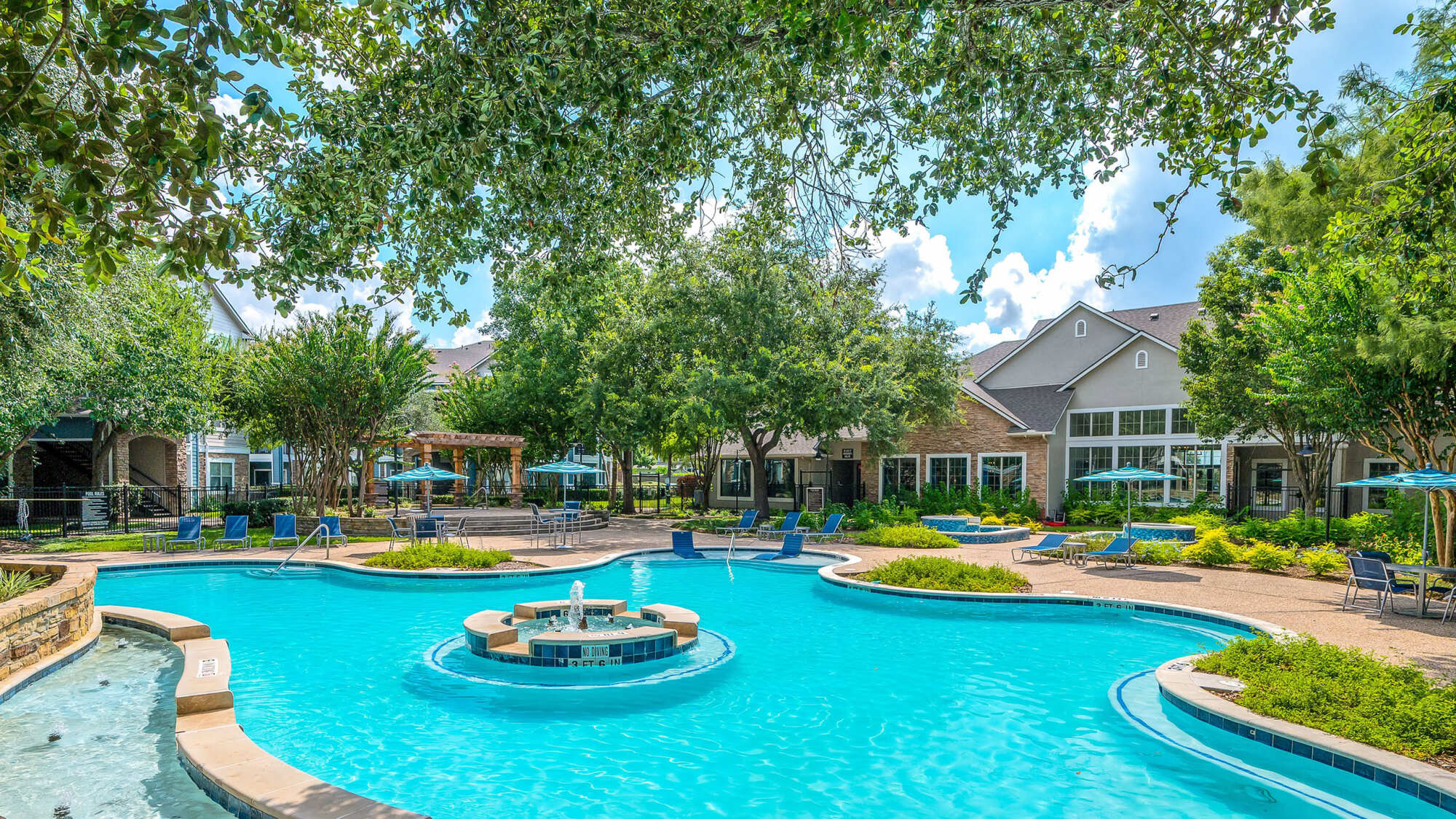 Lagoon-style community pool with a central fountain feature, surrounded by mature trees, lounge chairs, blue umbrellas, and the apartment clubhouse in the background.