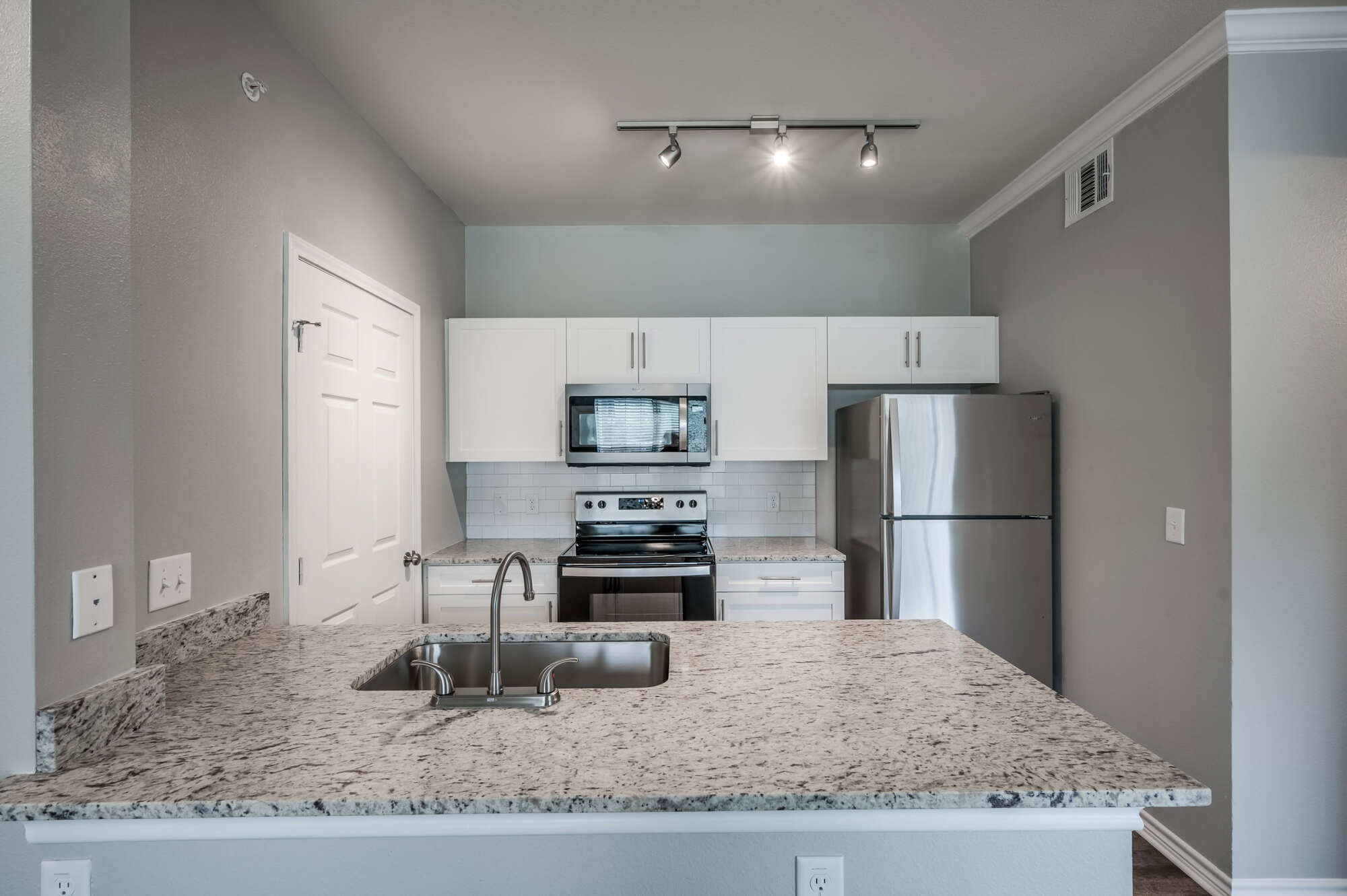 Apartment kitchen featuring white shaker cabinetry, granite-topped peninsula with stainless steel sink, stainless appliances, white subway tile backsplash, and modern track lighting.
