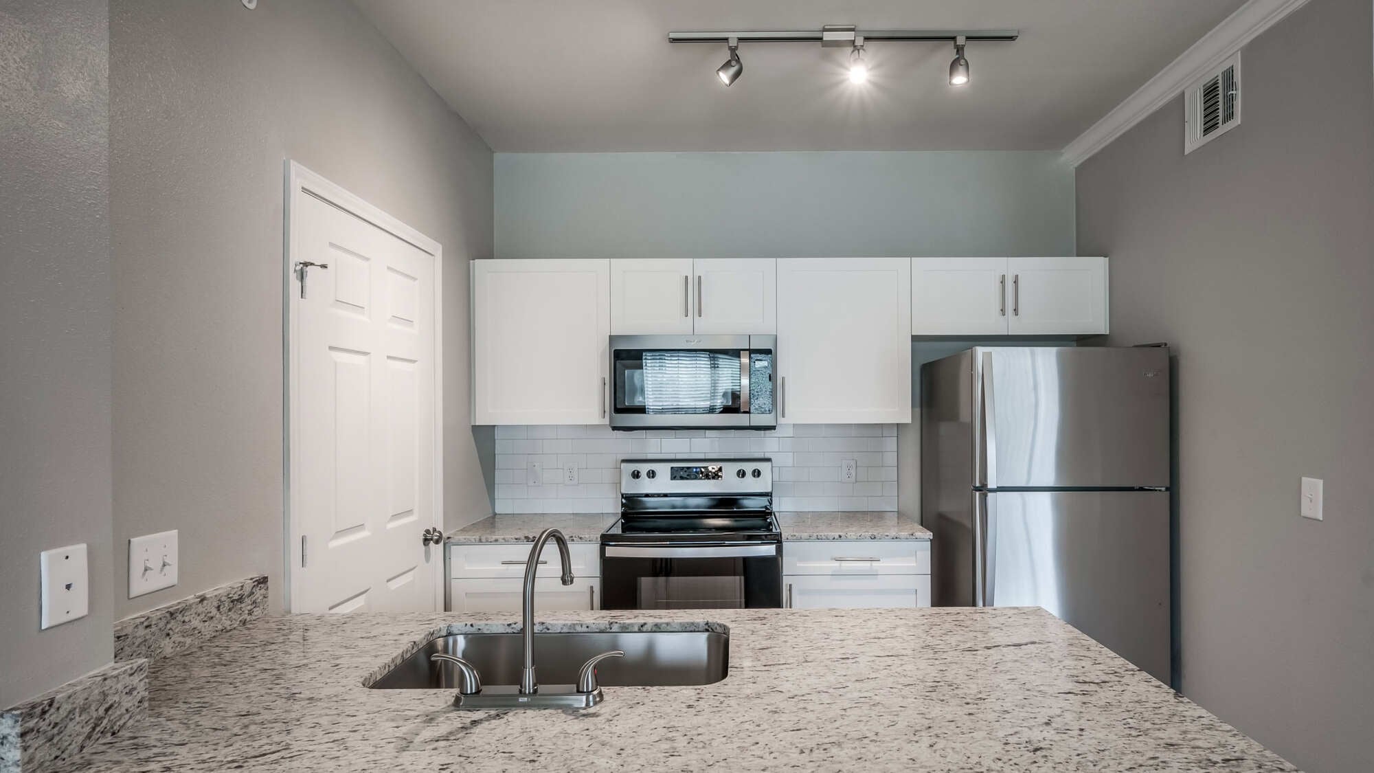 Clean apartment kitchen with white shaker cabinets, granite countertops, stainless steel appliances, white subway tile backsplash, modern track lighting, and a peninsula-style breakfast bar.