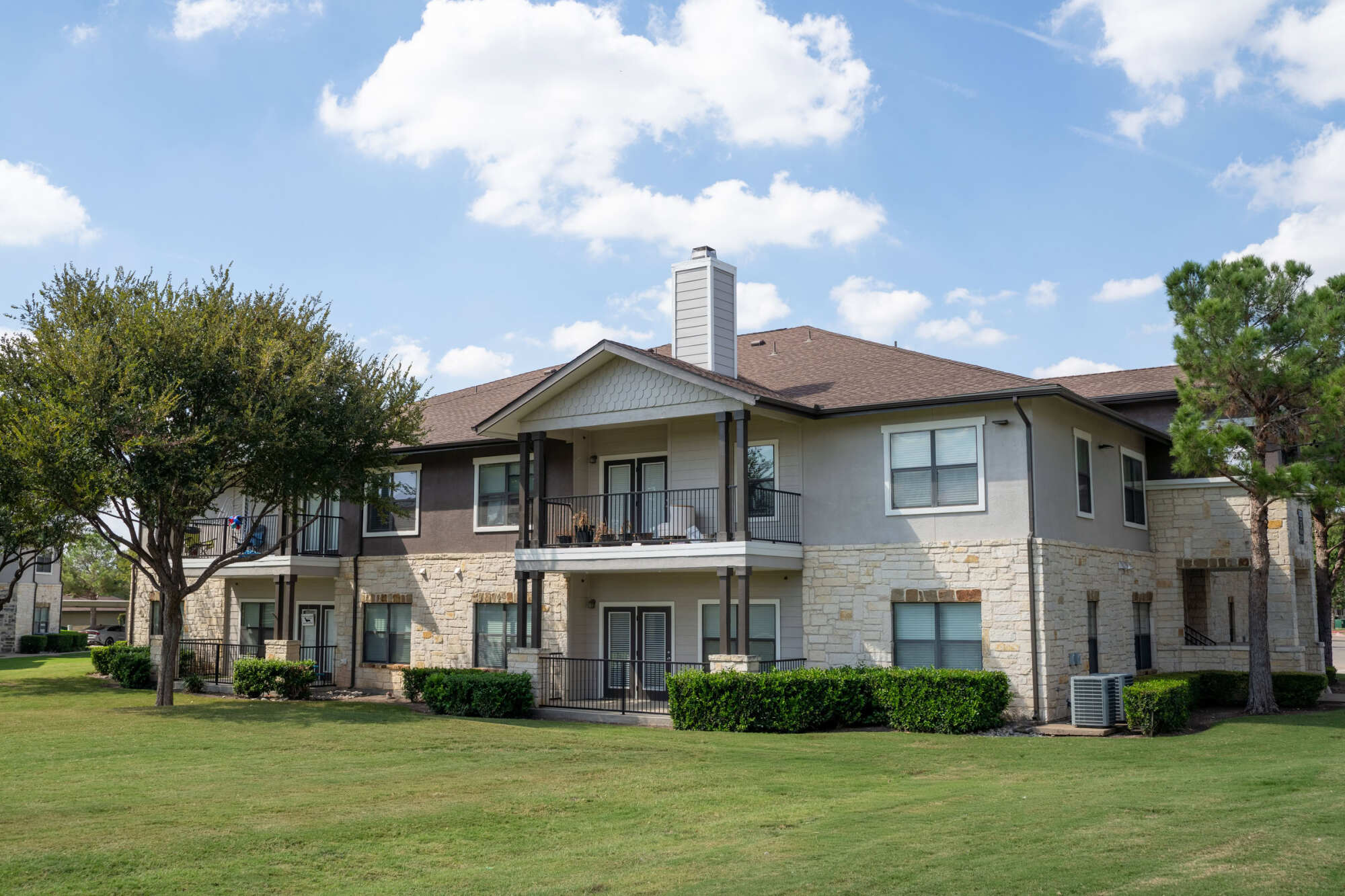 Two-story stucco and limestone apartment building with covered balconies, peaked dormer rooflines, manicured shrubs, mature trees, and a green lawn under a partly cloudy blue sky.