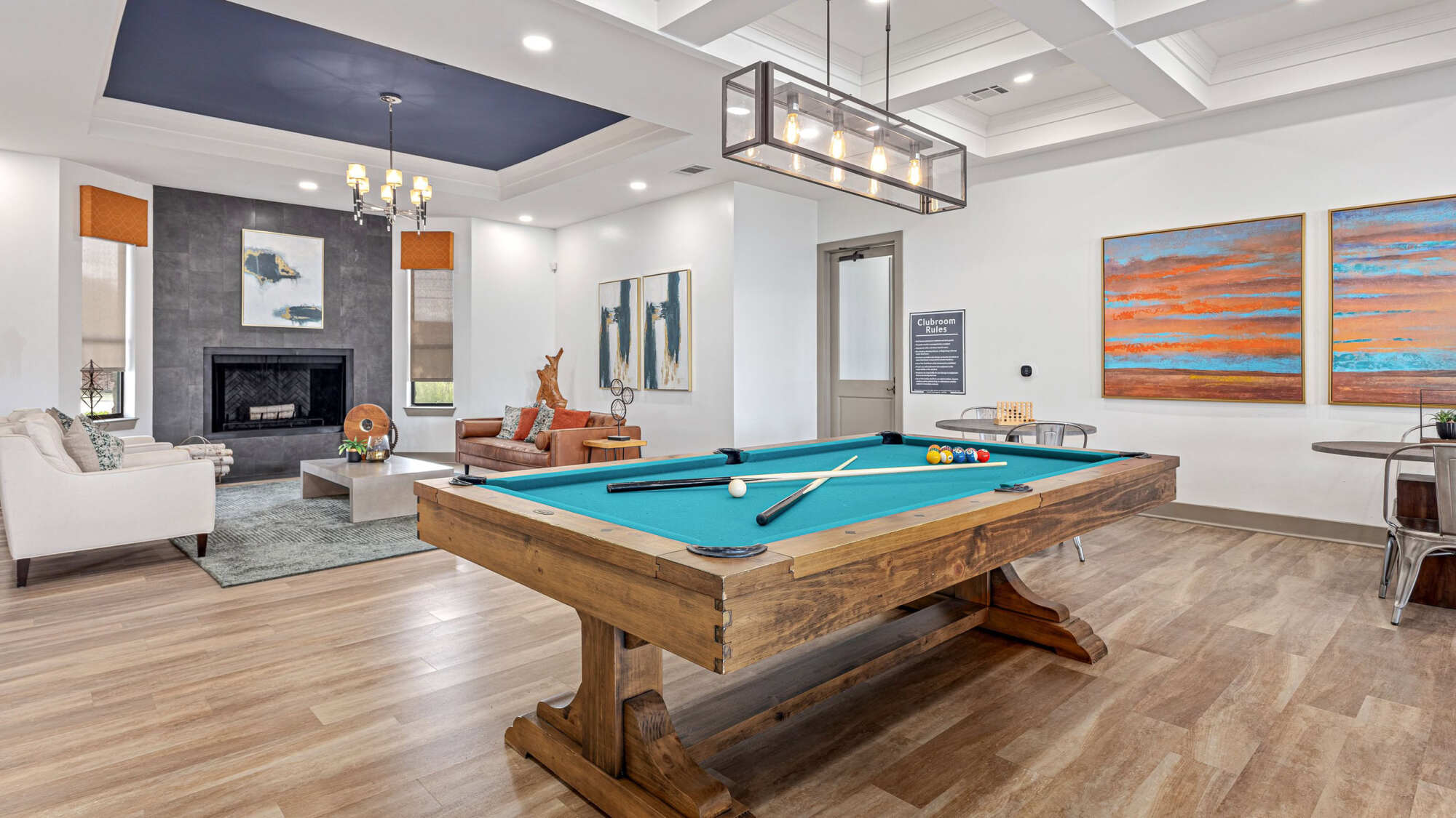 Stylish clubhouse game room with a teal-felt billiards table, modern linear chandelier, coffered ceiling, gray tile fireplace, lounge seating, and abstract sunset artwork on the walls.