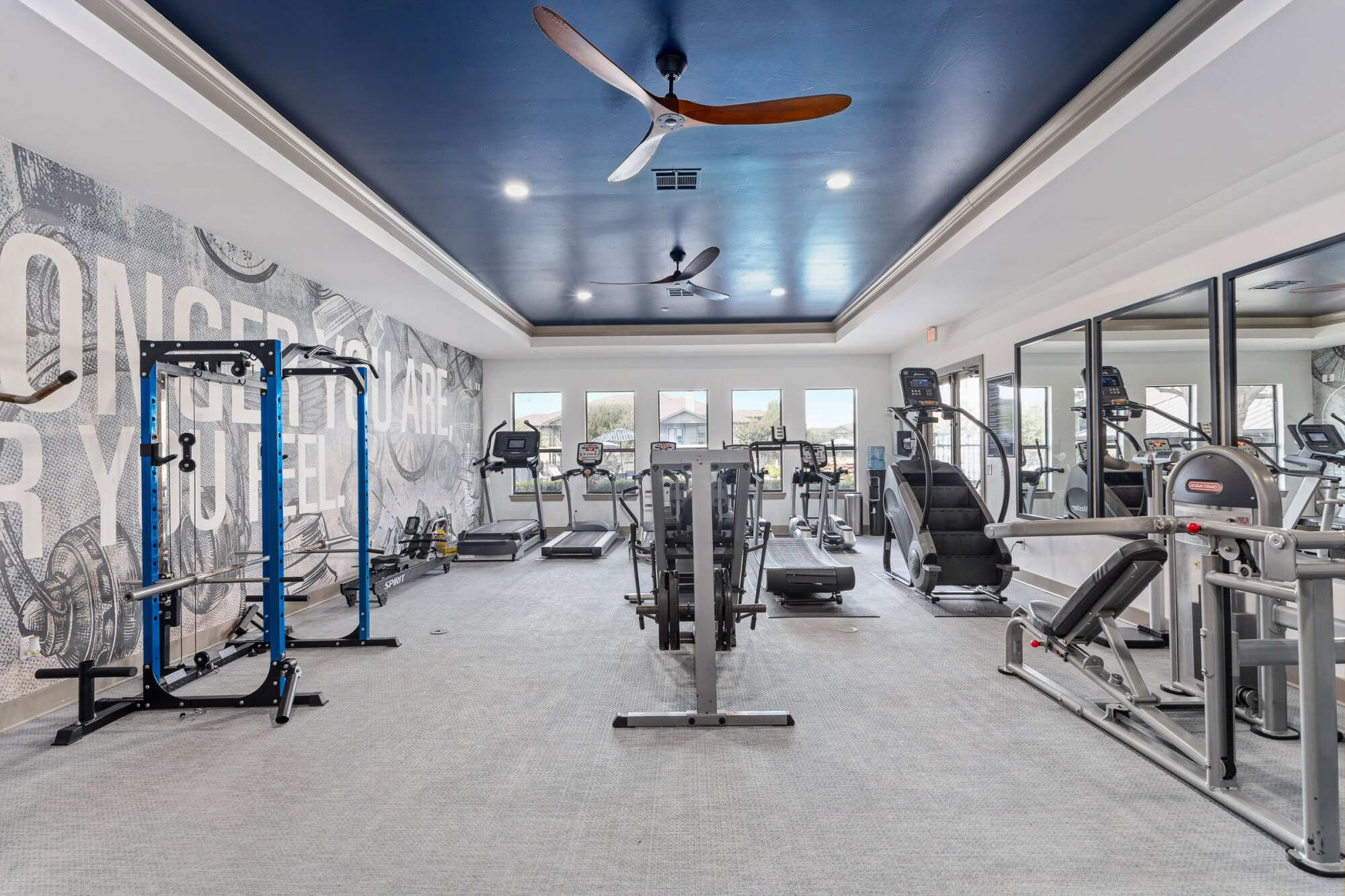Modern apartment gym with navy ceiling, wood-blade ceiling fans, treadmills, weight machines, a power rack, mirrored wall, and a graphic mural reading "Conquer Yourself."