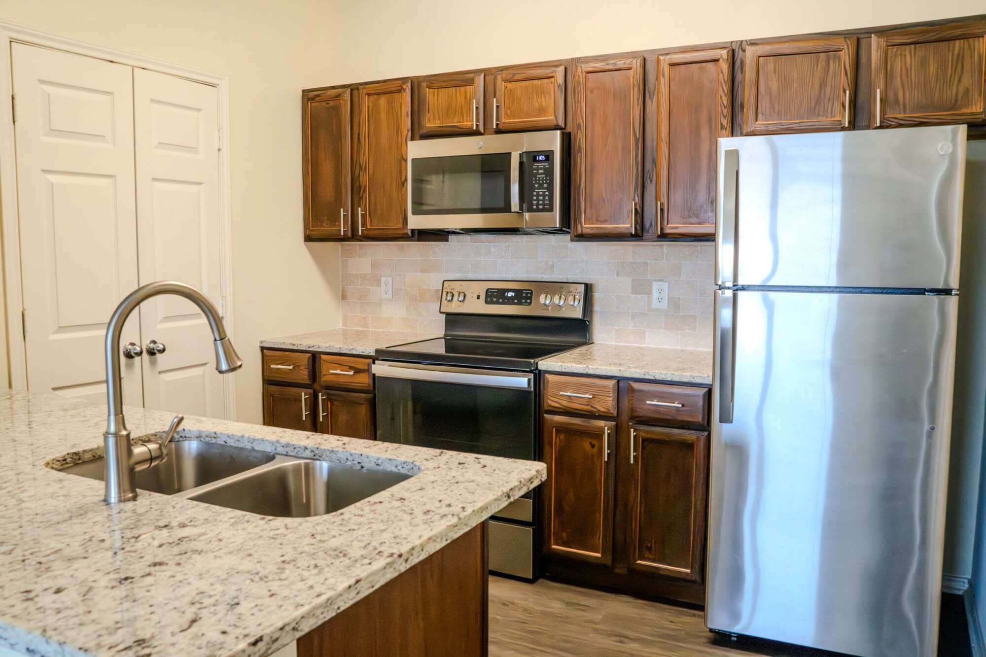 Apartment kitchen featuring warm wood cabinetry, granite countertops, stainless steel appliances, stone tile backsplash, and a peninsula with stainless steel sink and modern faucet.