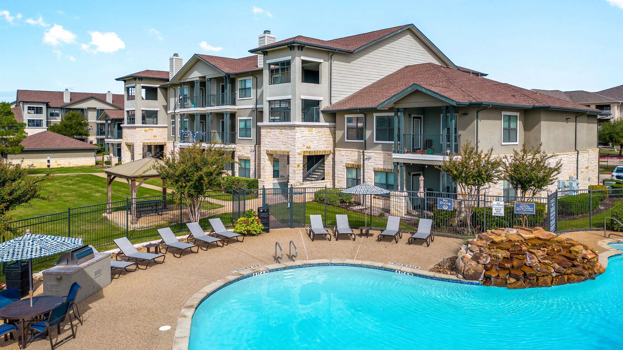Three-story apartment building with stone veneer accents and balconies, fronted by a curved swimming pool with lounge chairs, a stacked stone water feature, and shaded seating area.