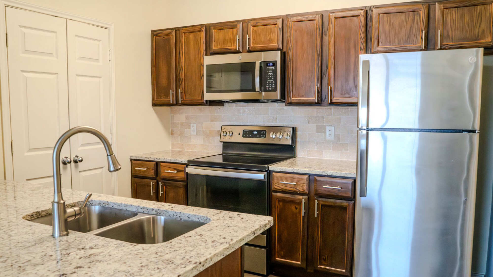Apartment kitchen featuring rich knotty wood cabinetry, granite countertops, stainless steel appliances, stone tile backsplash, and a peninsula with stainless steel double sink.