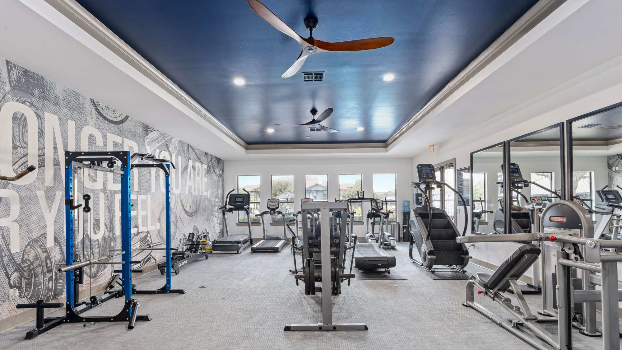 Modern fitness center with a navy blue tray ceiling, wood-blade ceiling fan, cardio machines, weight stations, a power rack, full mirrored wall, and graphic motivational wall art.