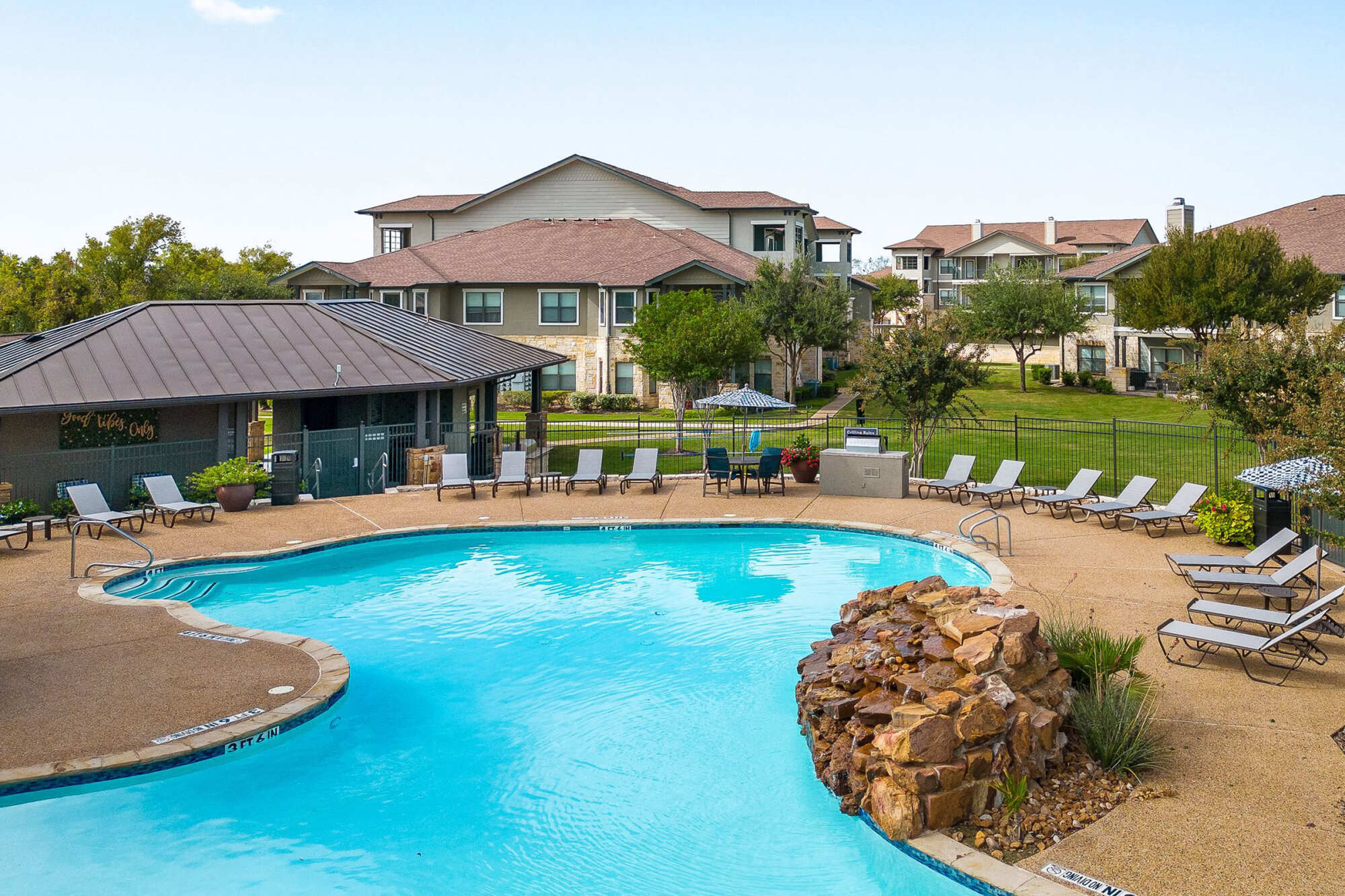Curved resort-style swimming pool with a rock waterfall feature, lounge chairs, paver decking, a pool cabana, and apartment buildings in the background under a hazy sky.