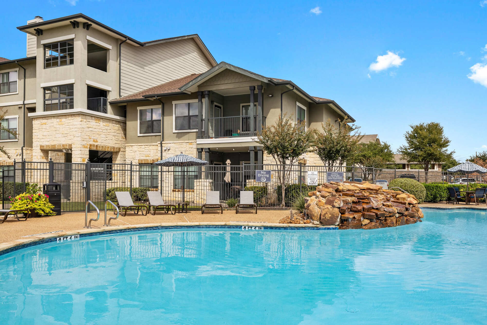 Inviting pool with crystal blue water, a stacked stone water feature, lounge chairs, manicured shrubs, and a two-story stucco apartment building with balconies overlooking the pool deck.