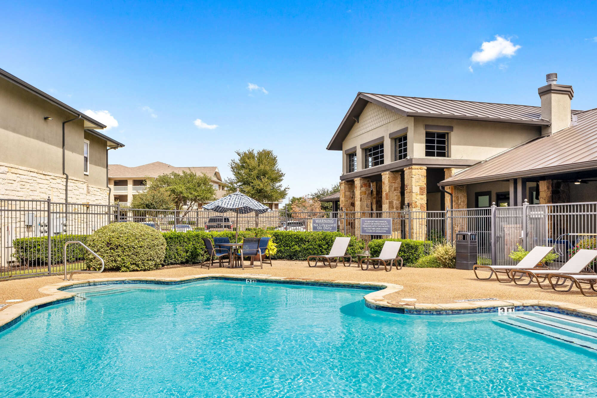 Sparkling community pool with lounge chairs, an outdoor seating area shaded by an umbrella, stone-accented clubhouse, peaked rooflines, and a wrought iron fence under a vivid blue sky.