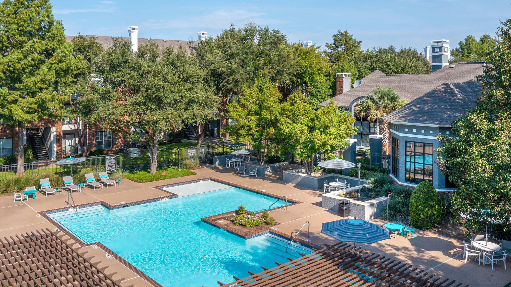 Aerial view of a resort-style community pool with crystal blue water, lounge chairs, blue umbrella shade, an elegant clubhouse with peaked roof, and mature shade trees.