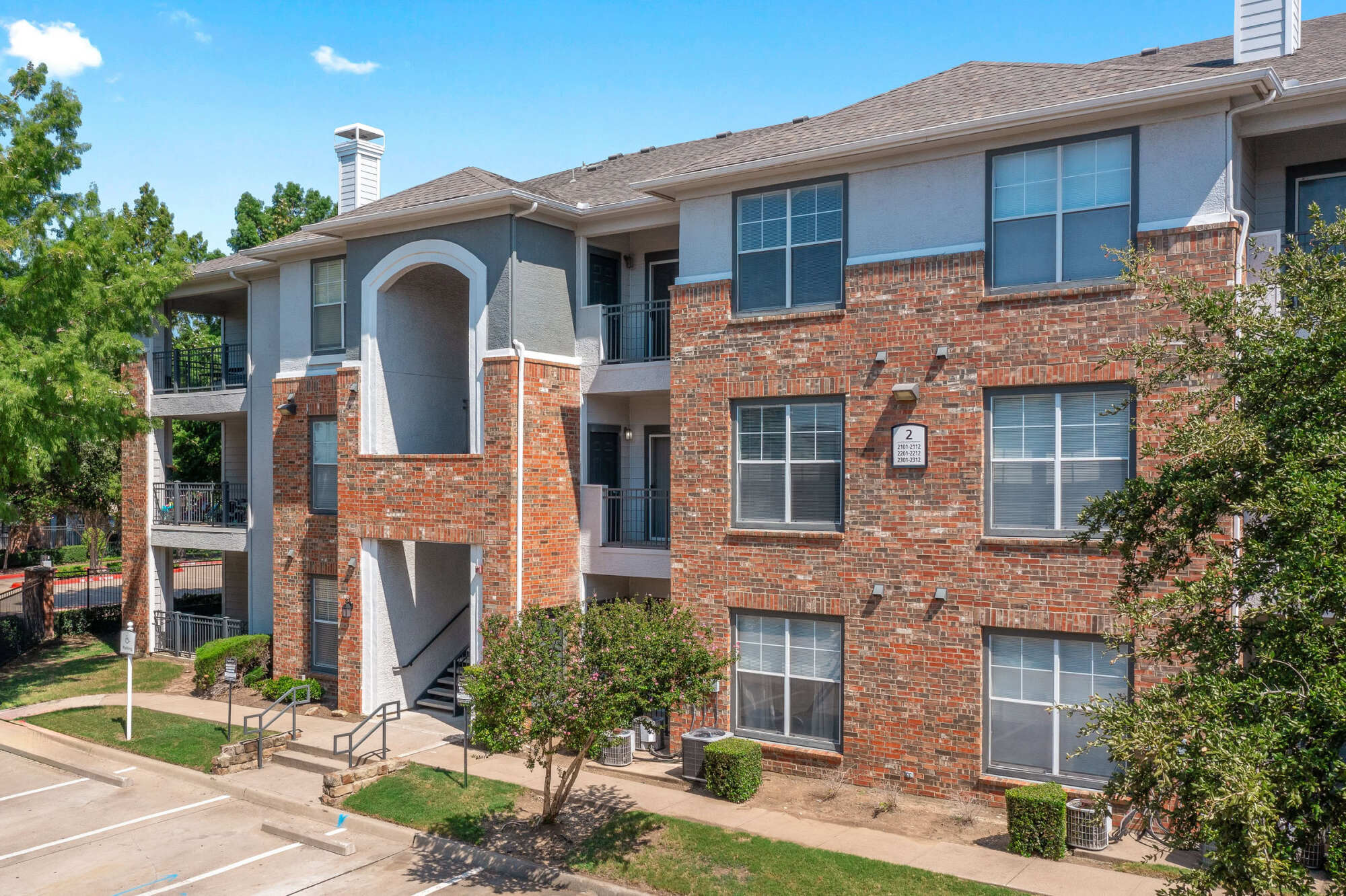 Three-story apartment building with red brick and stucco facade, arched architectural details, private balconies, mature trees, and exterior staircases under a bright sky.