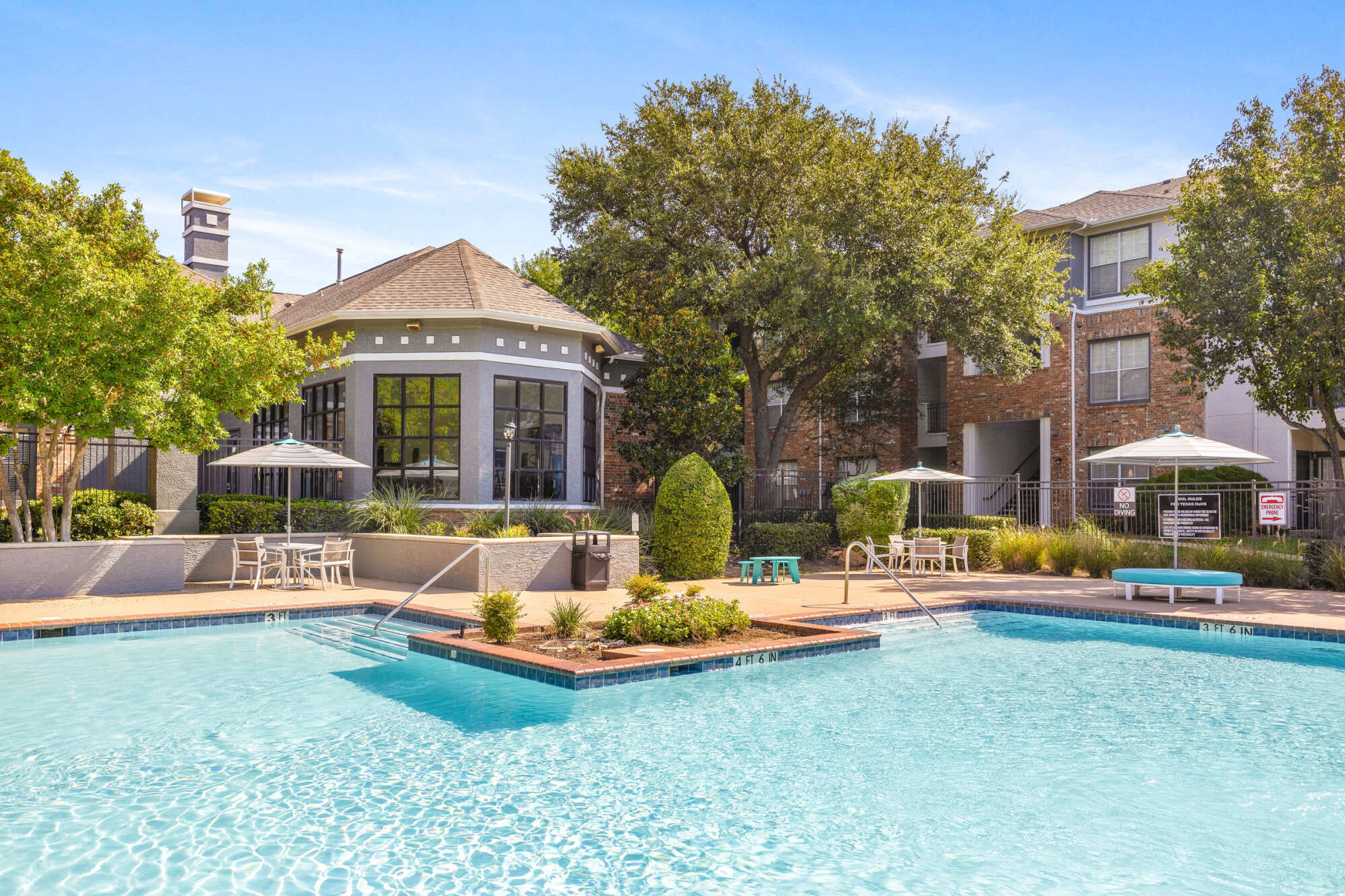Sparkling community pool flanked by an elegant gray clubhouse with arched windows, café seating with umbrellas, lush landscaping, and a vivid blue sky.