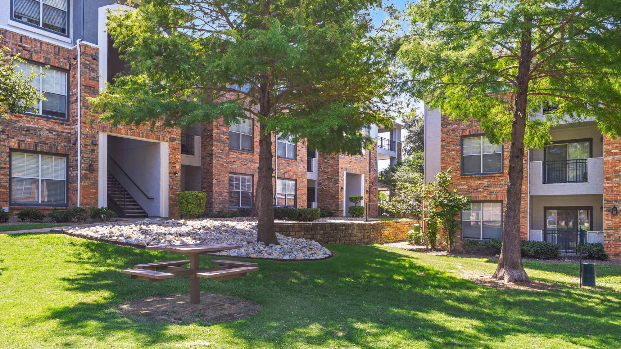 Brick apartment building exterior with mature cedar trees, river rock landscaping bed, a wooden picnic table, manicured green lawn, and an exterior staircase.
