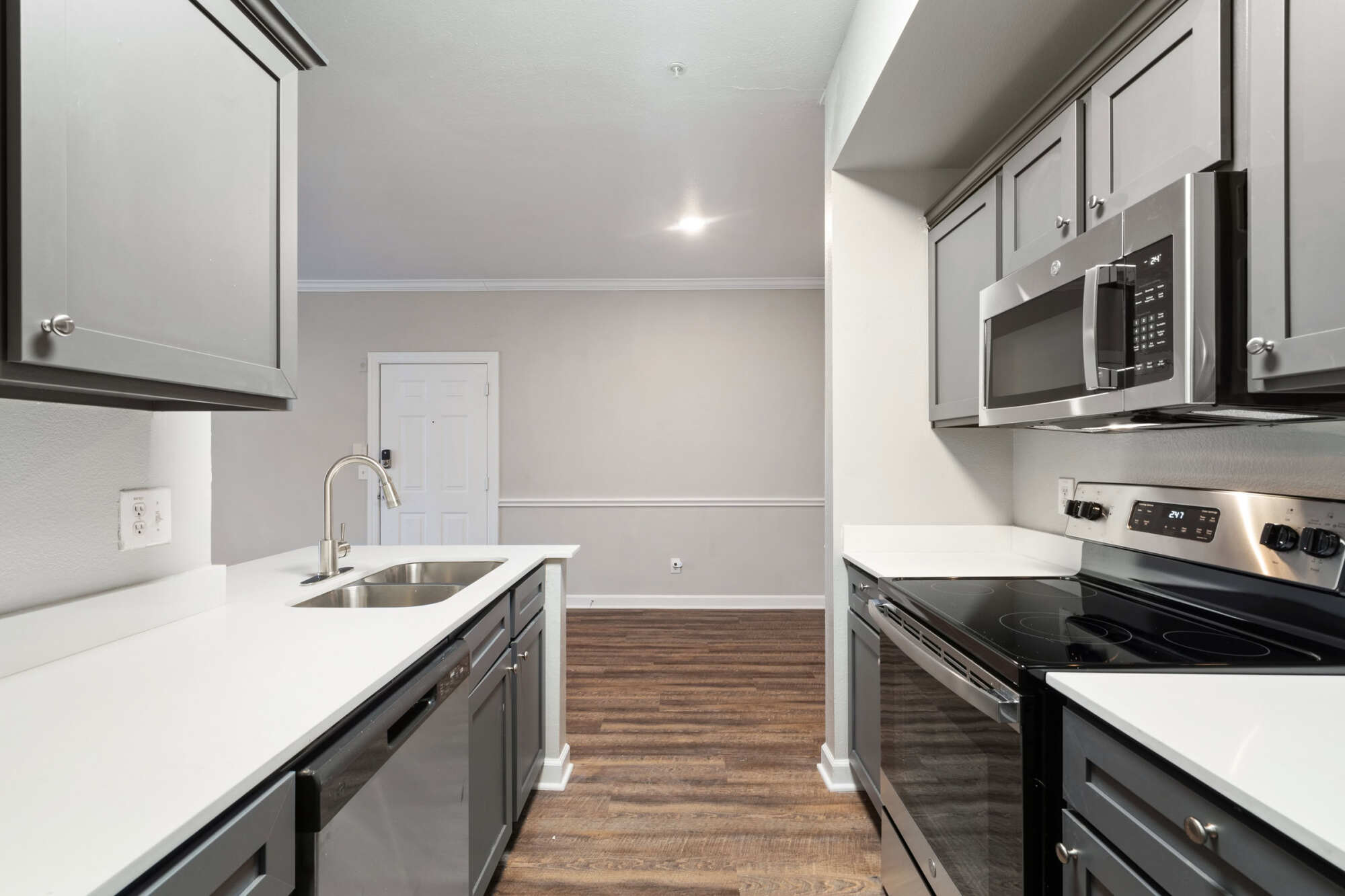 Open apartment kitchen with gray cabinetry, white quartz counters, stainless steel appliances, double basin sink, and a glimpse of the dining area with wood-look floors.