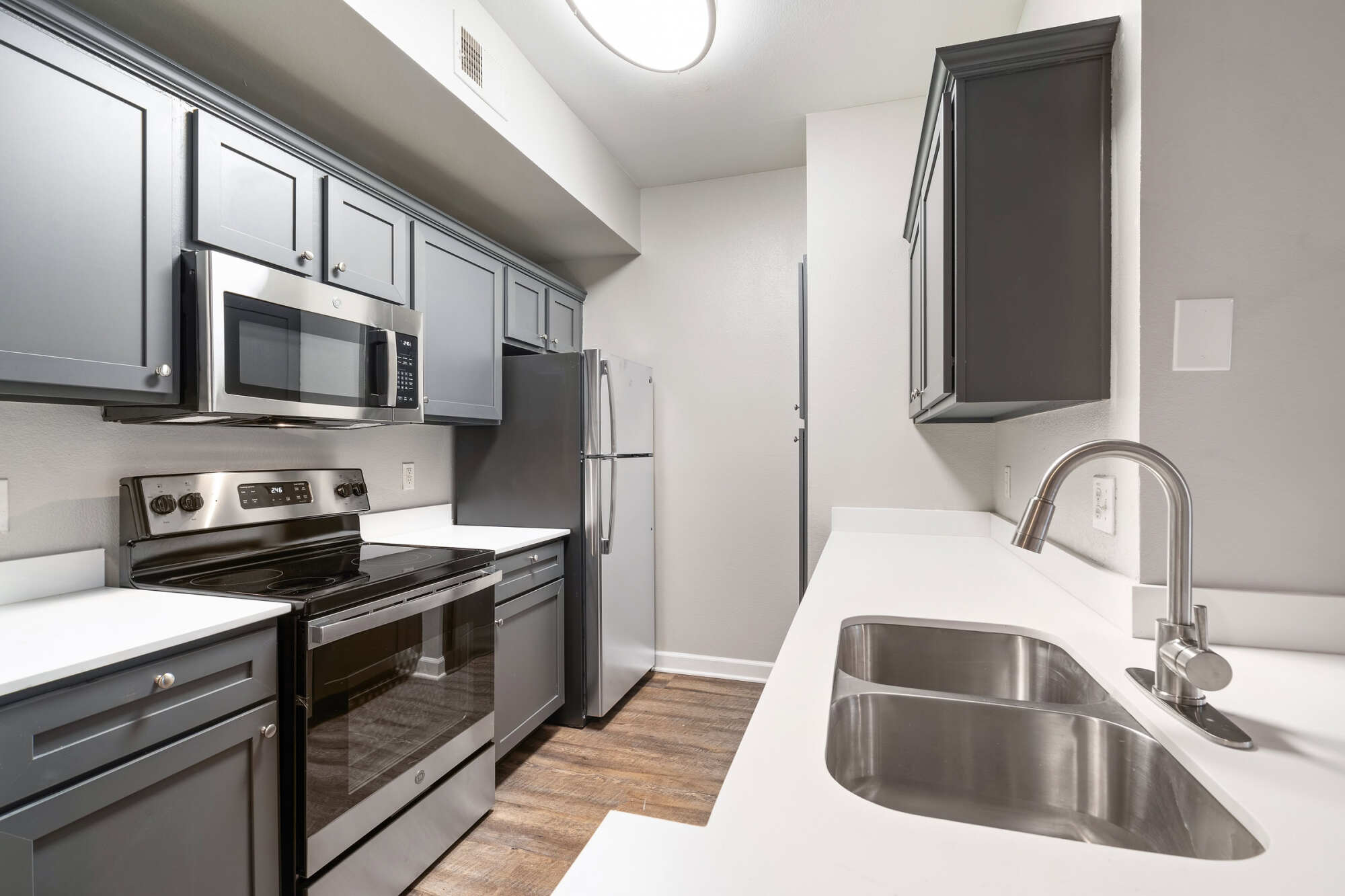 Galley apartment kitchen with gray shaker cabinets, white quartz countertops, stainless steel appliances, a deep double sink, and warm wood-look flooring.
