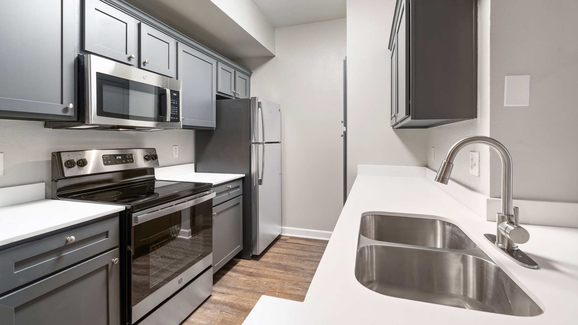 Modern galley kitchen featuring gray shaker cabinetry, white quartz countertops, stainless steel appliances, a deep double sink, and warm wood-look plank flooring.