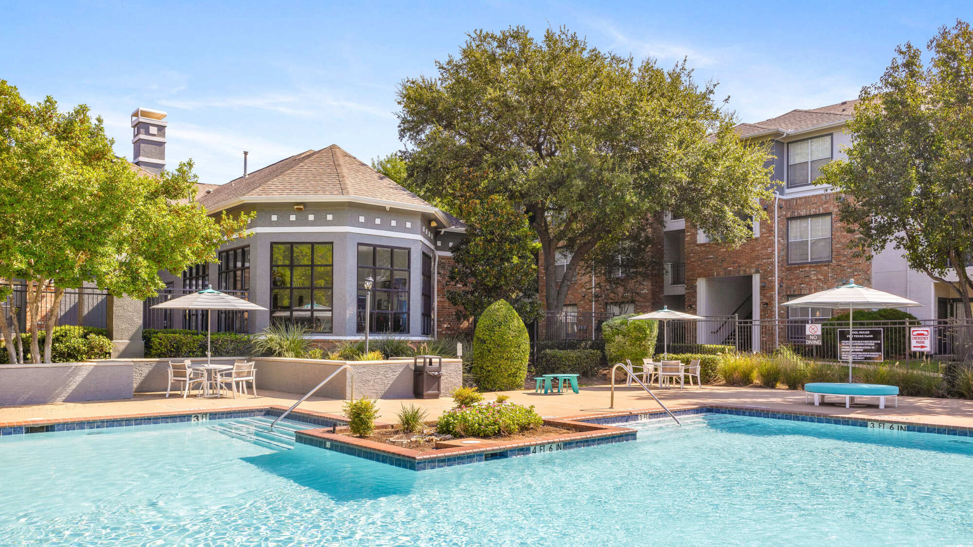 Inviting community pool with crystal blue water, a charcoal-gray clubhouse with arched windows, café tables with umbrellas, manicured shrubs, and shade trees.