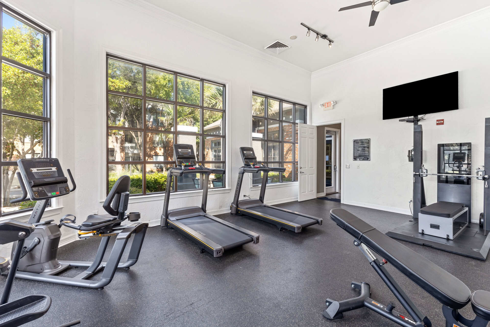 Apartment gym featuring a row of treadmills facing windows with a courtyard view, a recumbent bike, weight machine, and wall-mounted TV.
