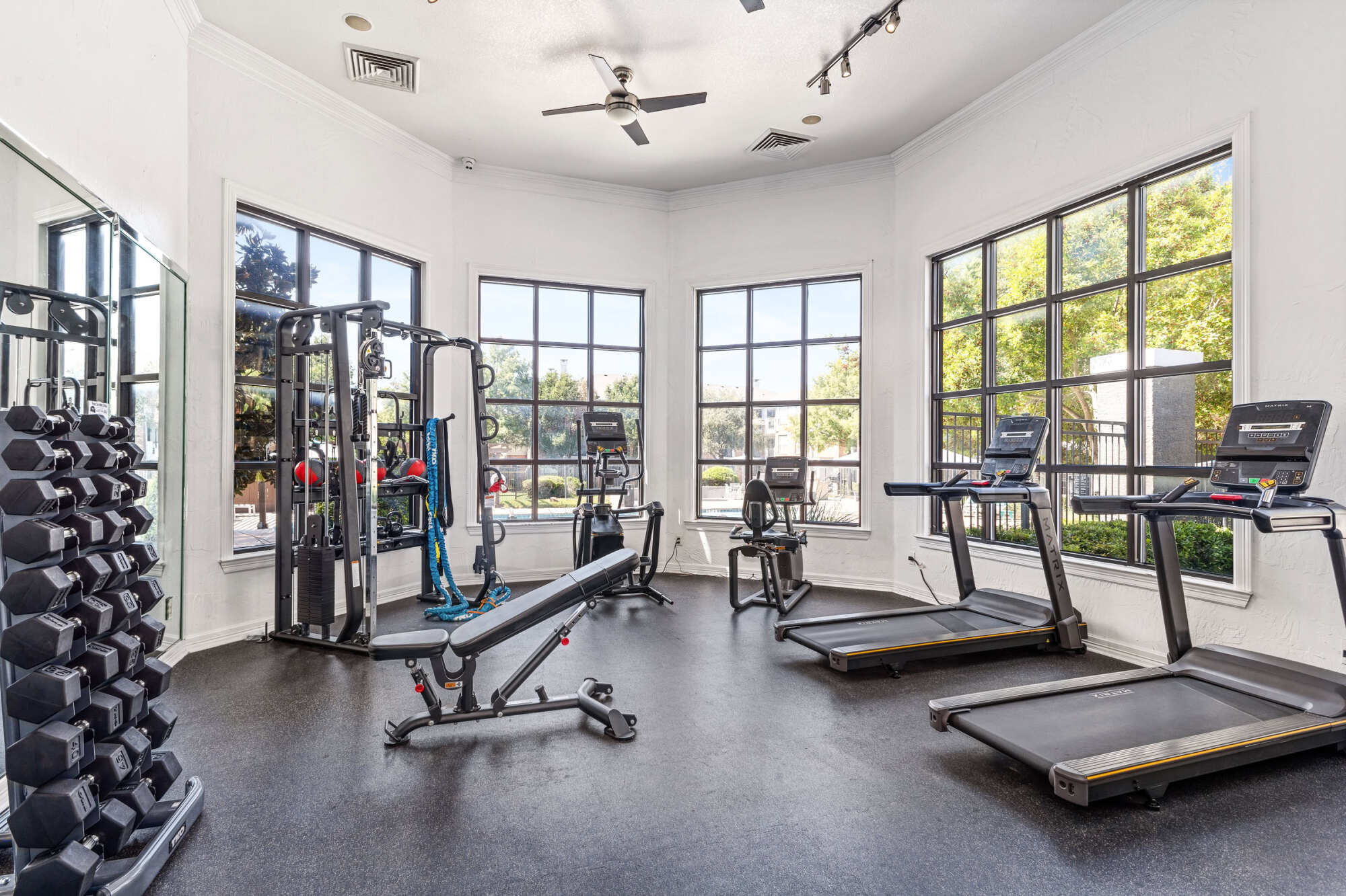 Bright fitness center with a dumbbell rack, multi-station cable machine, weight bench, treadmill, and large windows letting in natural light beneath a track-lit ceiling.