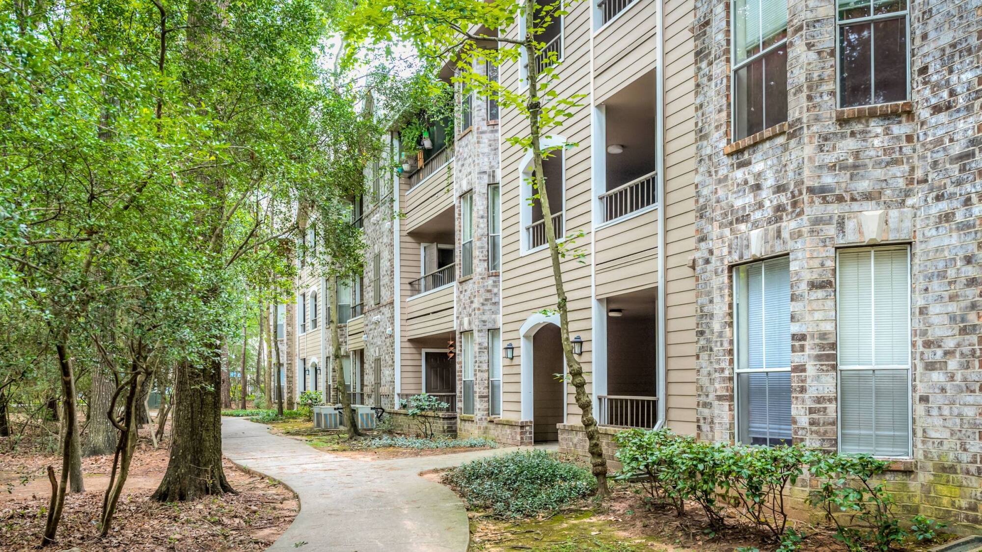 Shaded walking path beside a three-story Forest View apartment building in The Woodlands, TX, with private balconies and brick accents.