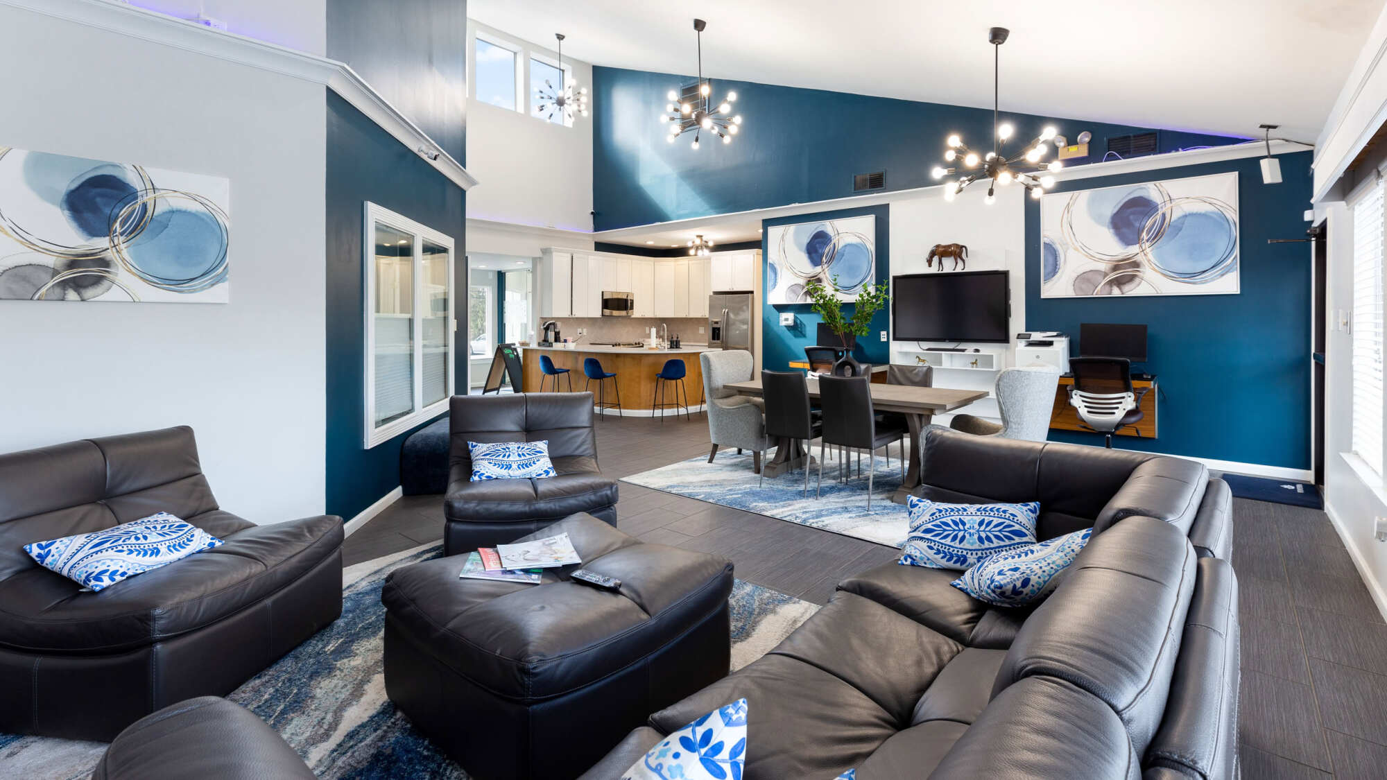 Spacious apartment clubhouse living room with vaulted ceilings, a navy blue accent wall, modern chandeliers, black leather sofas, and an open-concept kitchen and dining area.