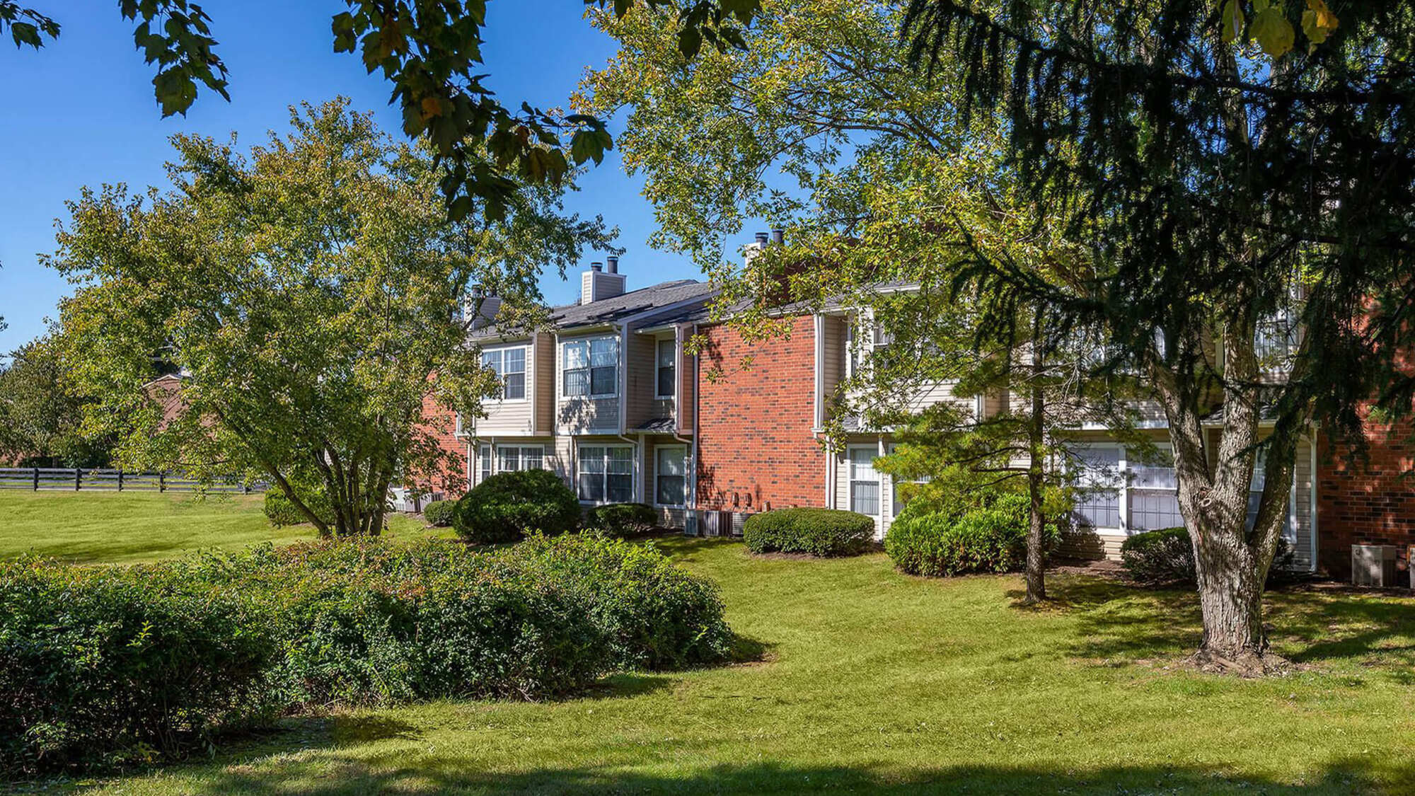 Landscaped grounds at Steeplechase Apartments framed by lush green trees and shrubs, with a brick apartment building visible in the background.