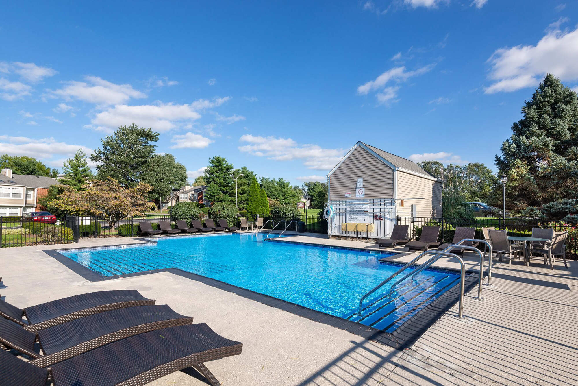 Resort-style outdoor pool with crystal-clear water, wicker lounge chairs, patio seating, and a tan pool house surrounded by trees on a sunny day.
