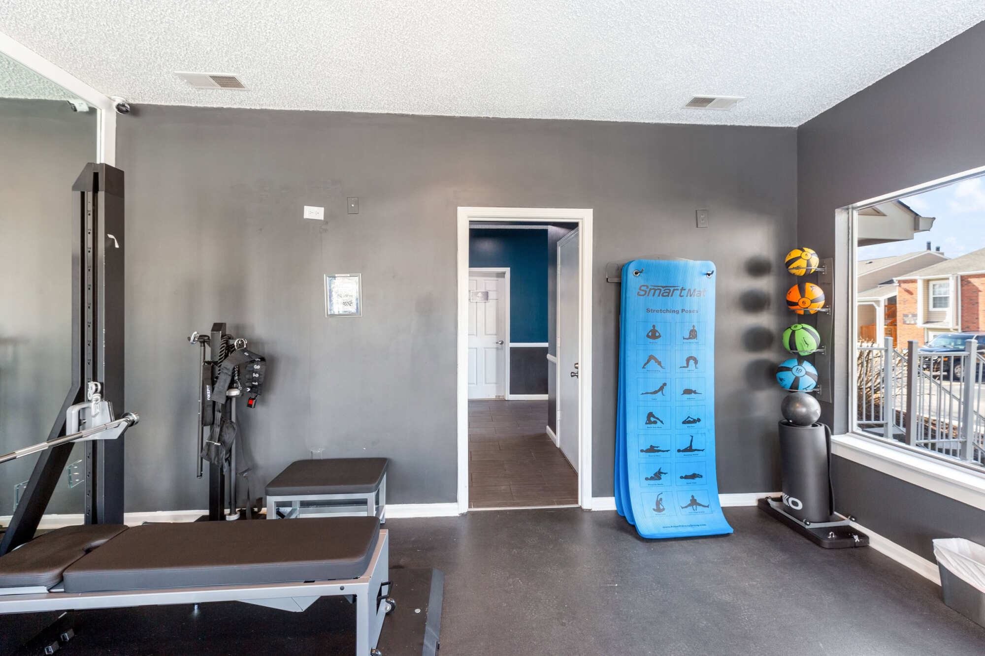 Fitness center area with a strength-training machine, weight bench, stability balls, and a yoga mat against gray walls, with a window overlooking the grounds.