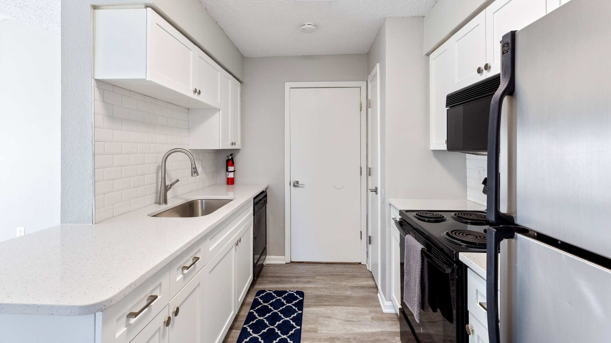 Renovated galley kitchen featuring white cabinetry, quartz countertops, white subway tile backsplash, stainless steel refrigerator, and black range and dishwasher.