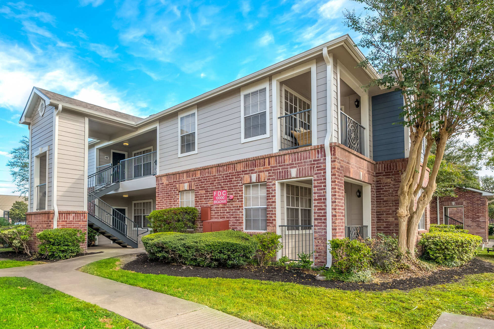 Exterior of a two-story apartment building with gray siding and brick accents, an exterior staircase, wrought-iron balconies, and landscaped shrubs under a blue sky.