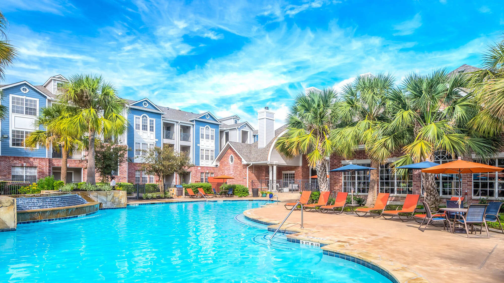 Sparkling blue swimming pool surrounded by a paved deck with palm trees, orange lounge chairs, umbrellas, and blue-trimmed apartment buildings under a bright sky.