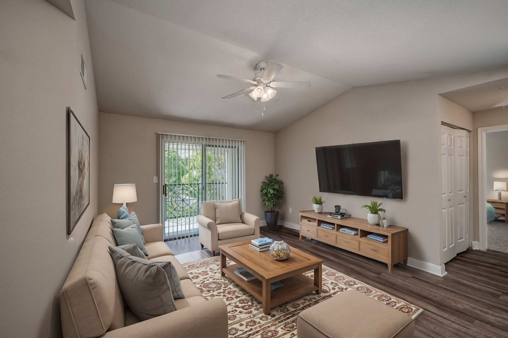 Cozy furnished living room with vaulted ceiling and ceiling fan, beige sofa and armchair, wall-mounted TV, wood media console, and a sliding glass door to a balcony.