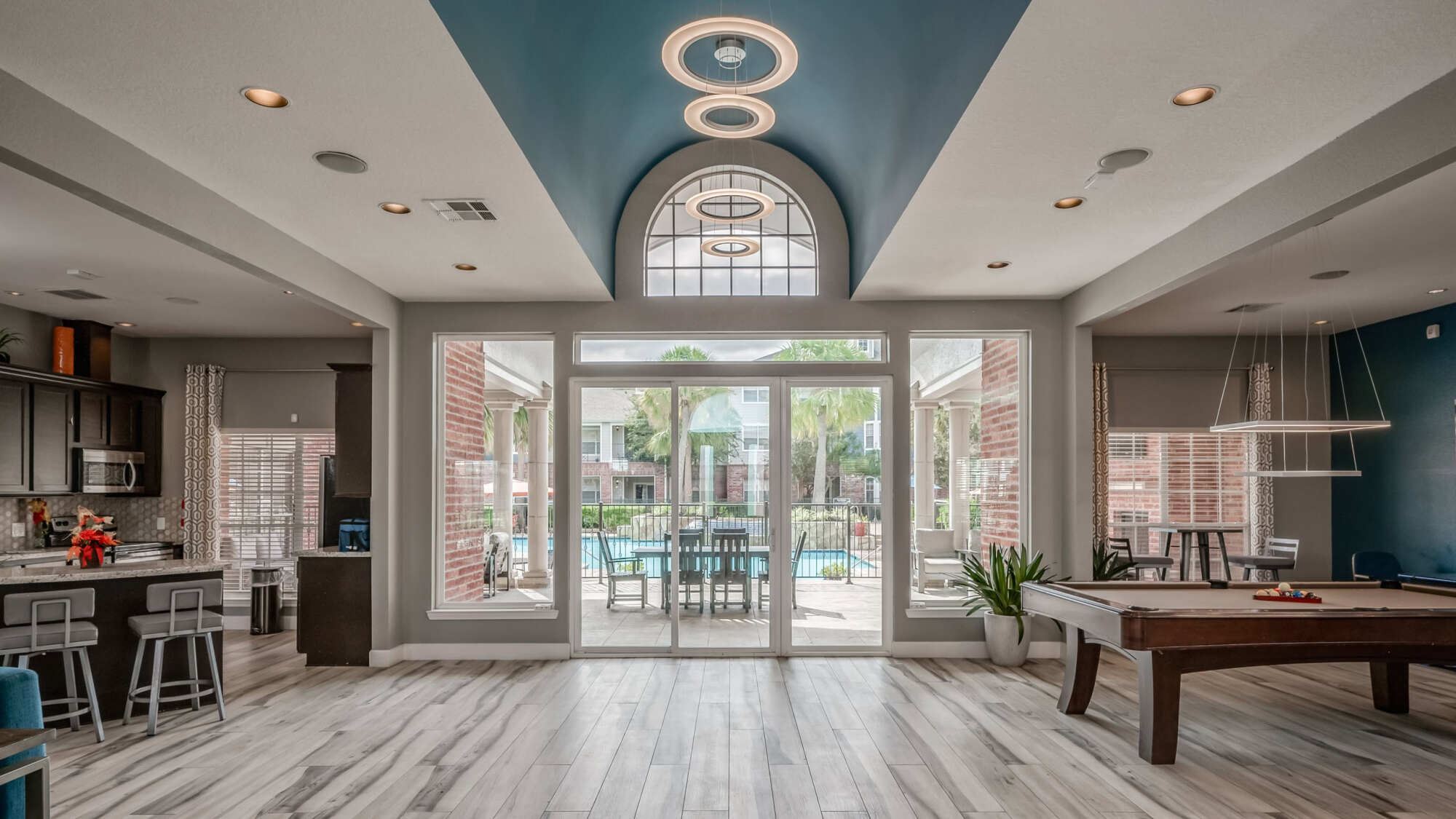 Open clubhouse interior with a blue accent ceiling, arched window, recessed lighting, a kitchen and bar area, a billiards table, and glass doors leading to the pool.