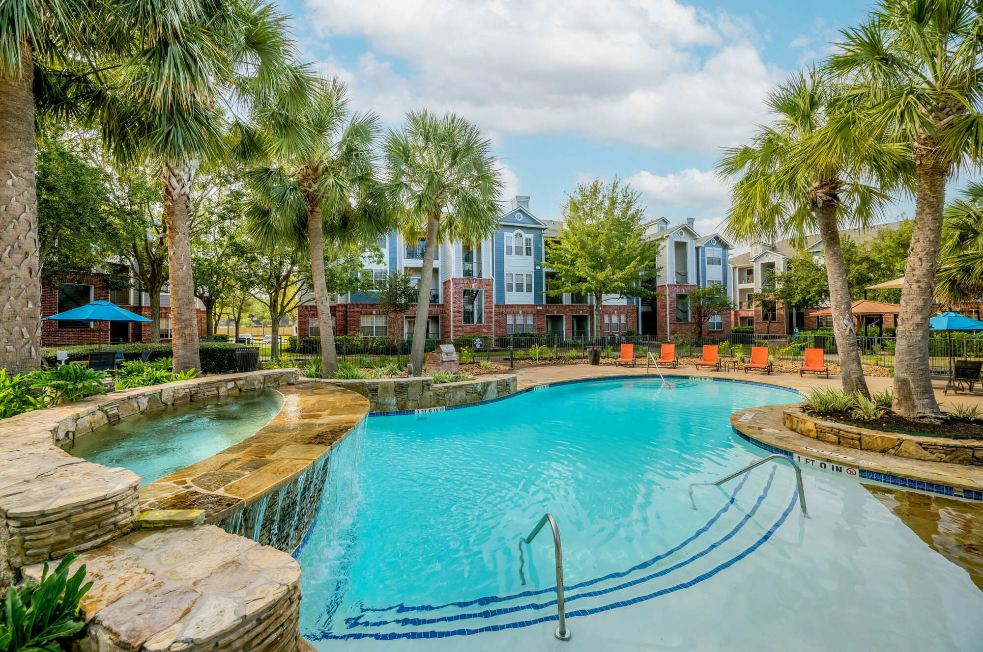 Curving resort-style pool with a stone waterfall spa, framed by palm trees, blue and orange umbrellas, lounge chairs, and brick apartment buildings.
